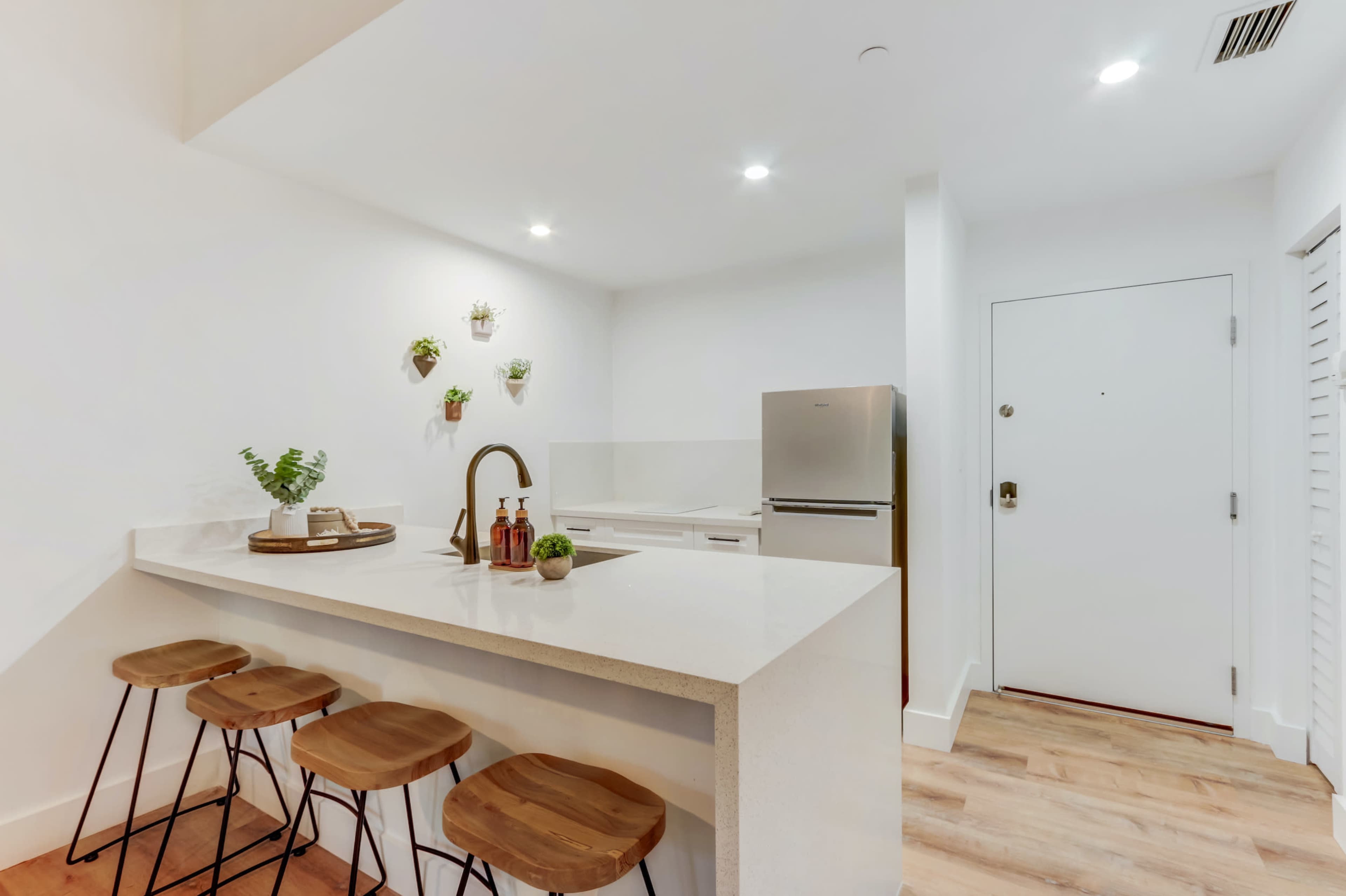 A modern kitchen with a white countertop island and three wooden stools, featuring a stainless steel refrigerator and a white door.