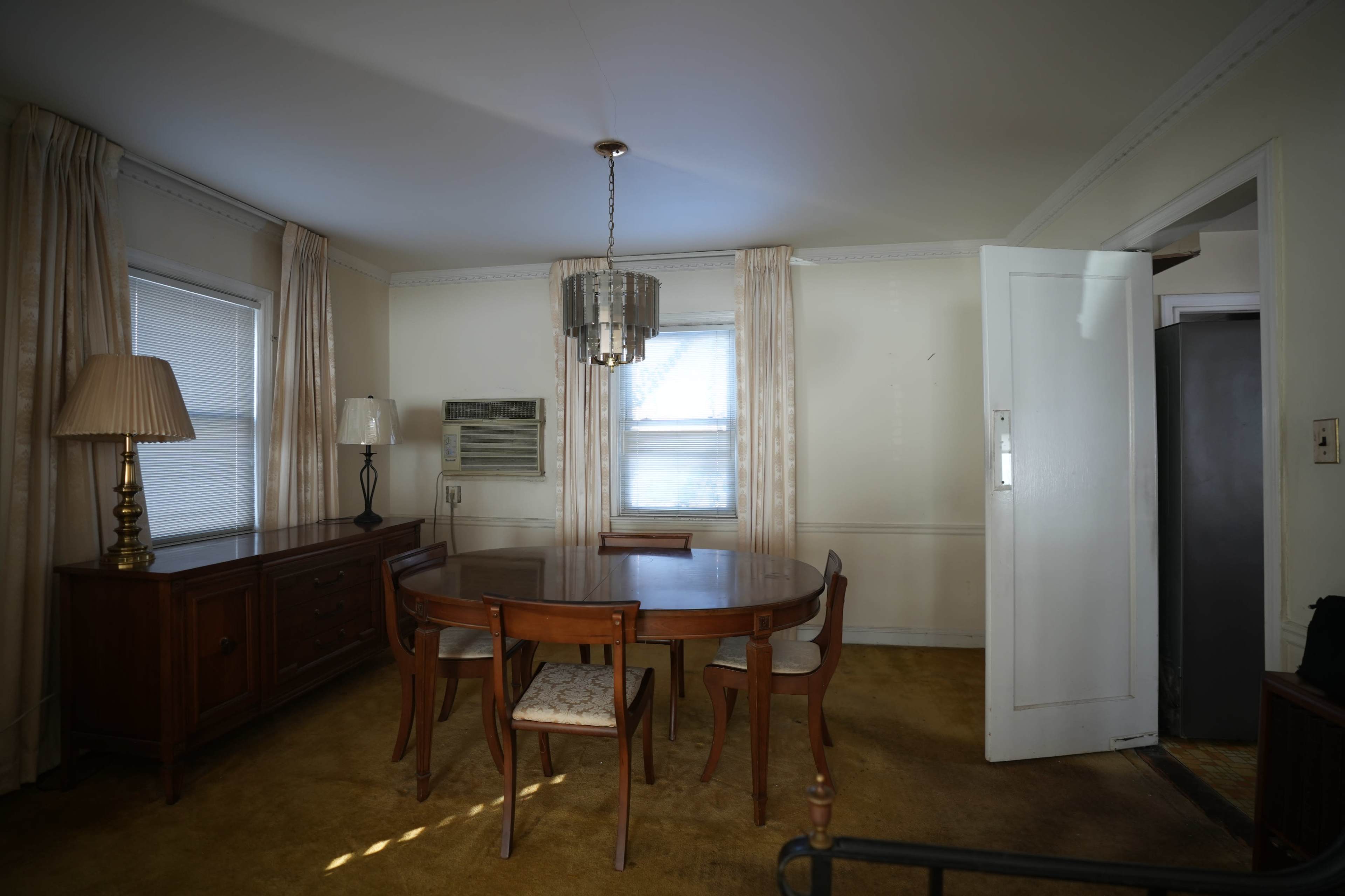 The image shows a dining area with a round table and several chairs, a lamp on a console table, and a window with sheer curtains, all situated in a room with beige carpeting.