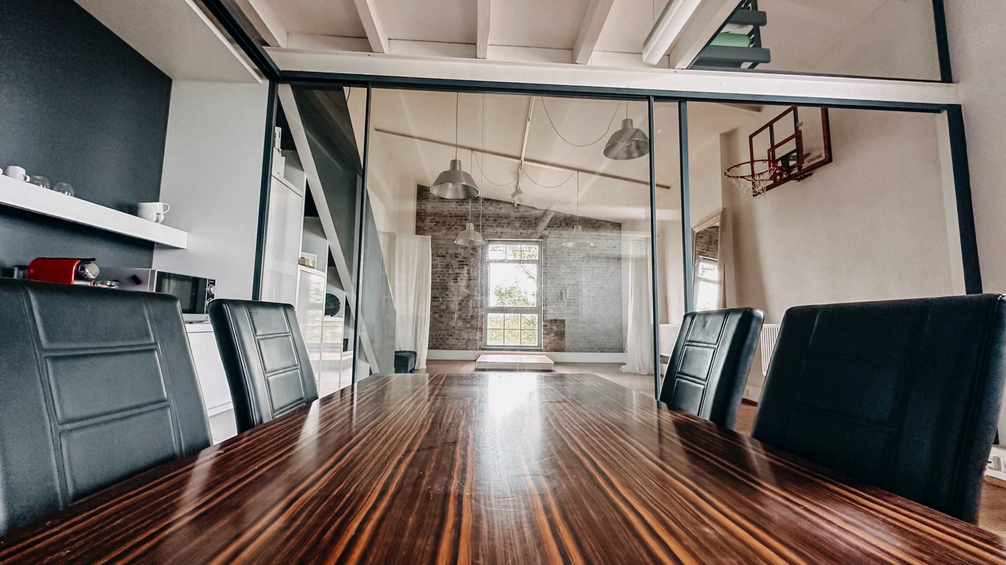 A modern conference room features a sleek wooden table and black chairs, with large glass walls revealing a brick-walled area that includes a basketball hoop.