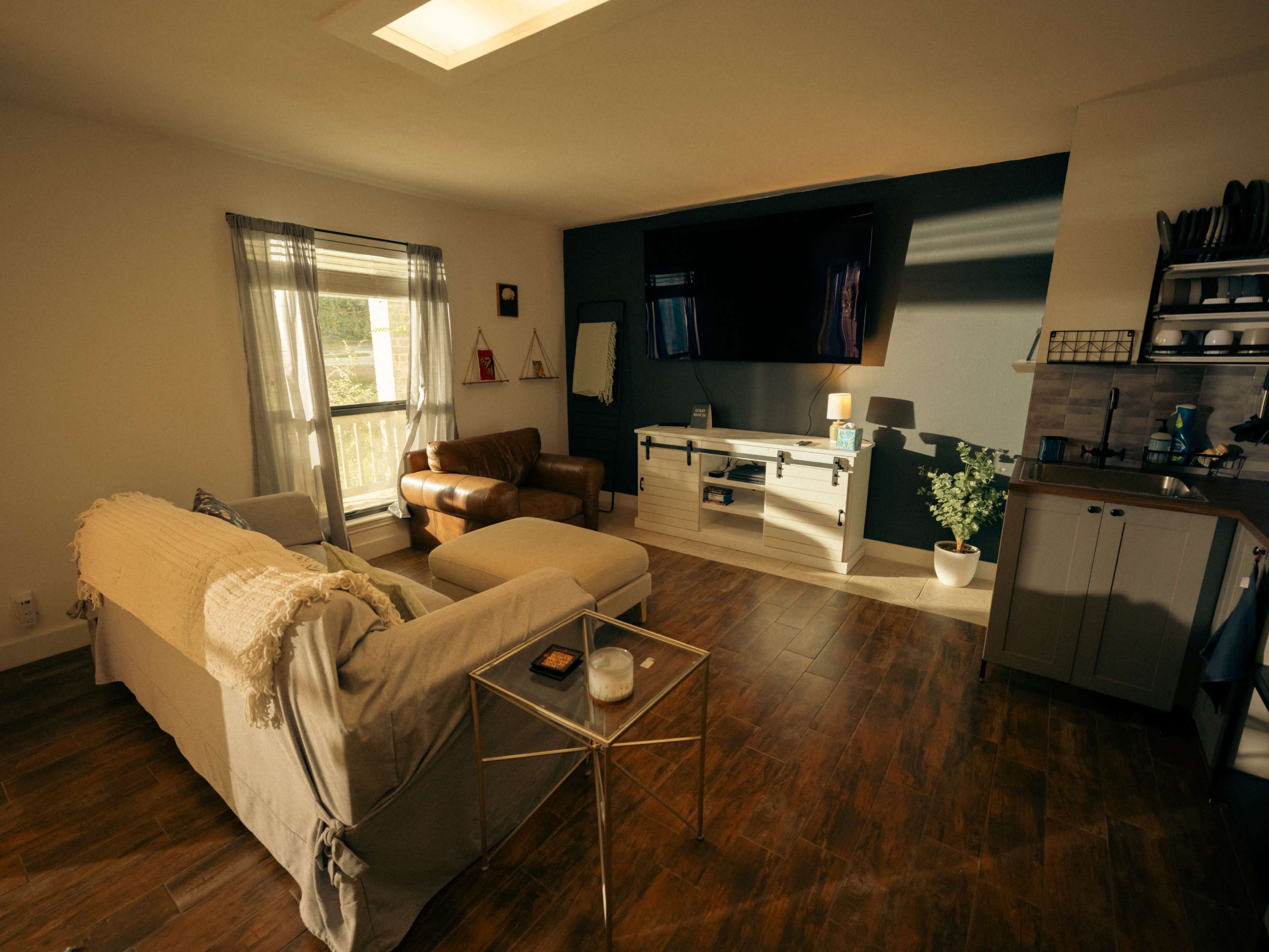The image shows a modern, cozy living room featuring a sofa, an armchair, a coffee table, and a television mounted on the wall.