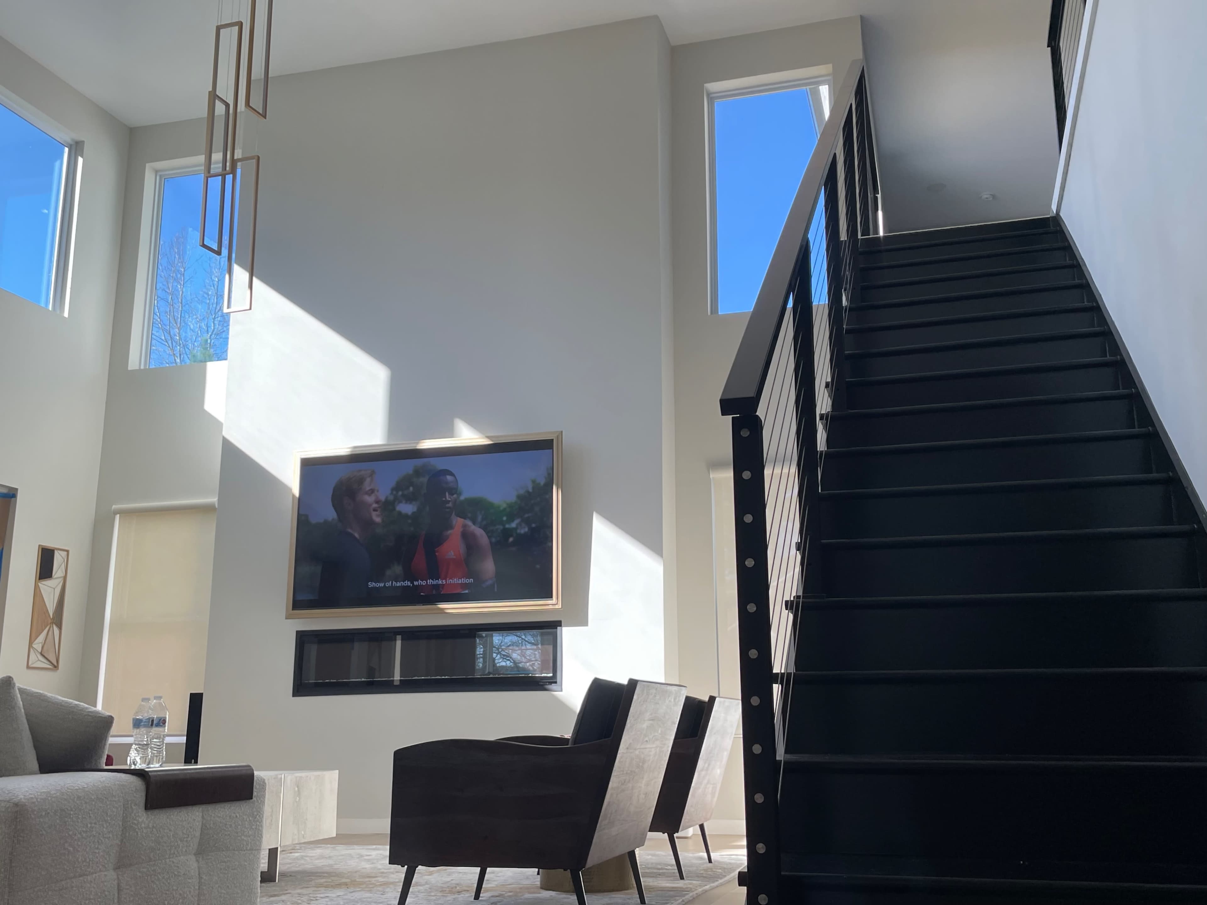 A modern living room with a large flat-screen TV mounted on the wall, two armchairs, and a staircase leading to an upper level.