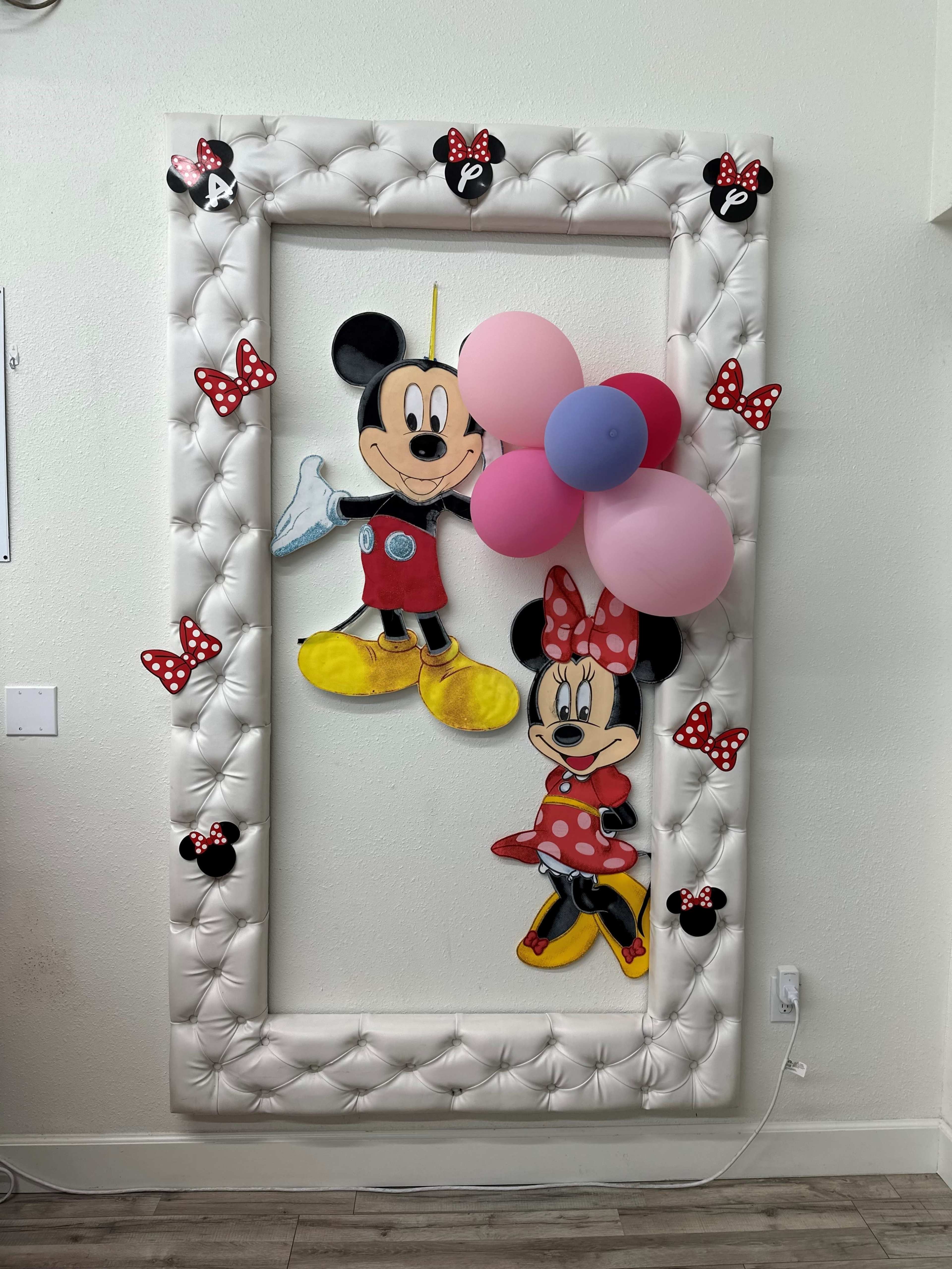 A large, padded white frame decorated with cartoon cutouts of Mickey and Minnie Mouse, along with colorful balloons and polka-dotted bows.