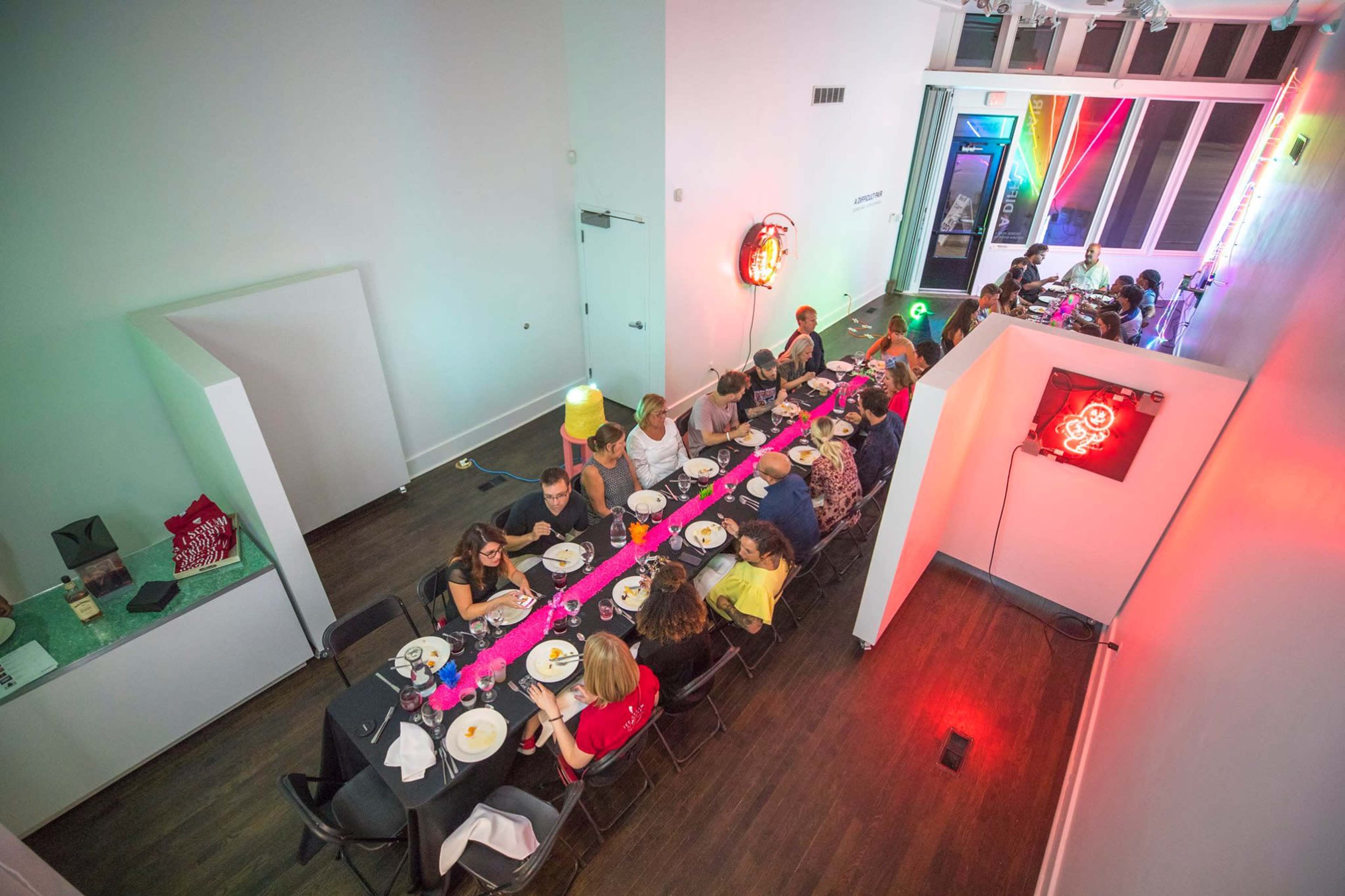 A large group of people is seated at a long black table, enjoying a meal in a brightly lit modern space with vibrant decorations and neon signs.