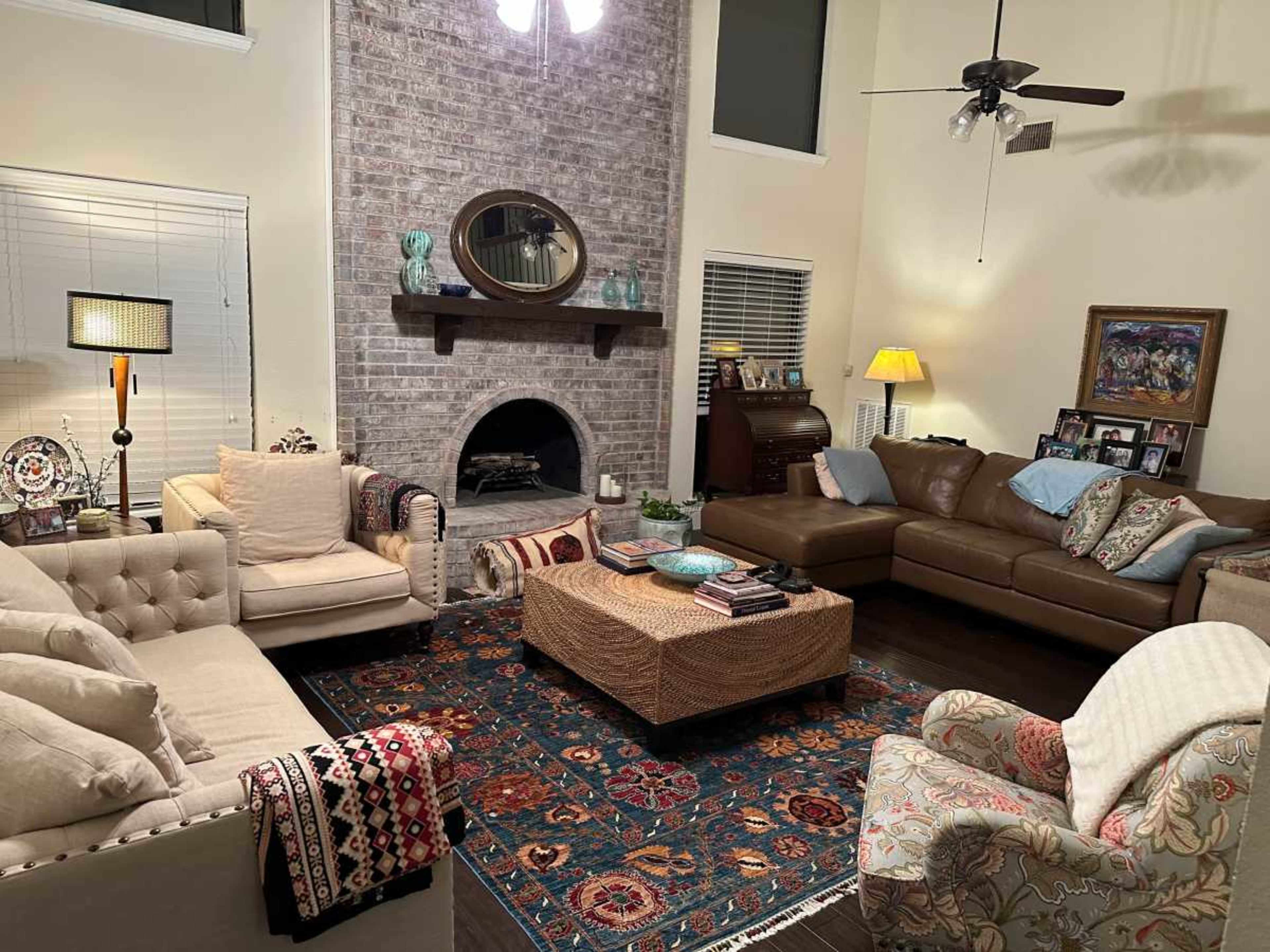 A cozy living room features two sofas, an armchair, and an ottoman arranged around a central area rug, with a brick fireplace and decorative items on the shelves.