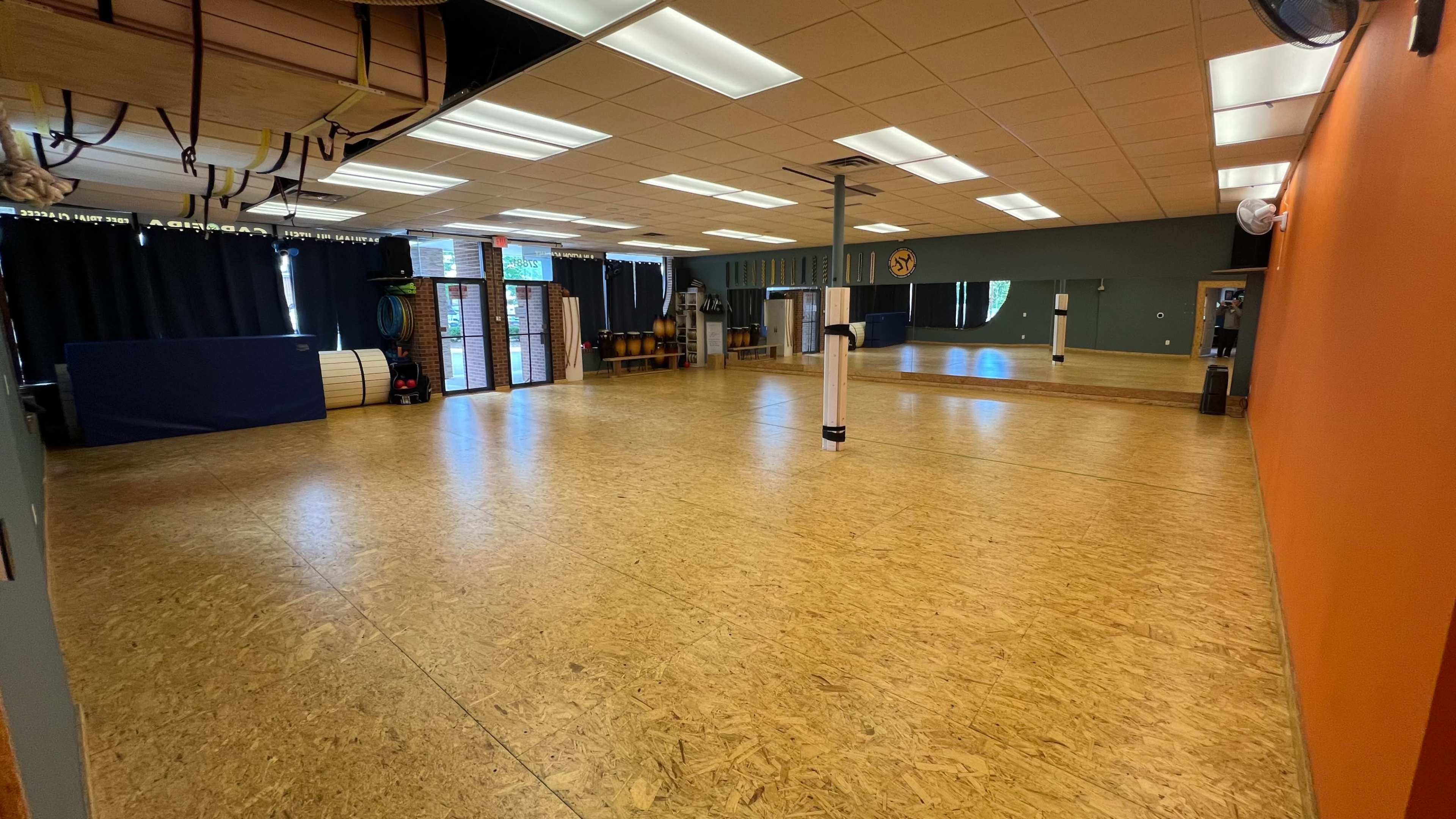 An empty fitness studio with wooden flooring, mirrors, and exercise equipment along the walls.