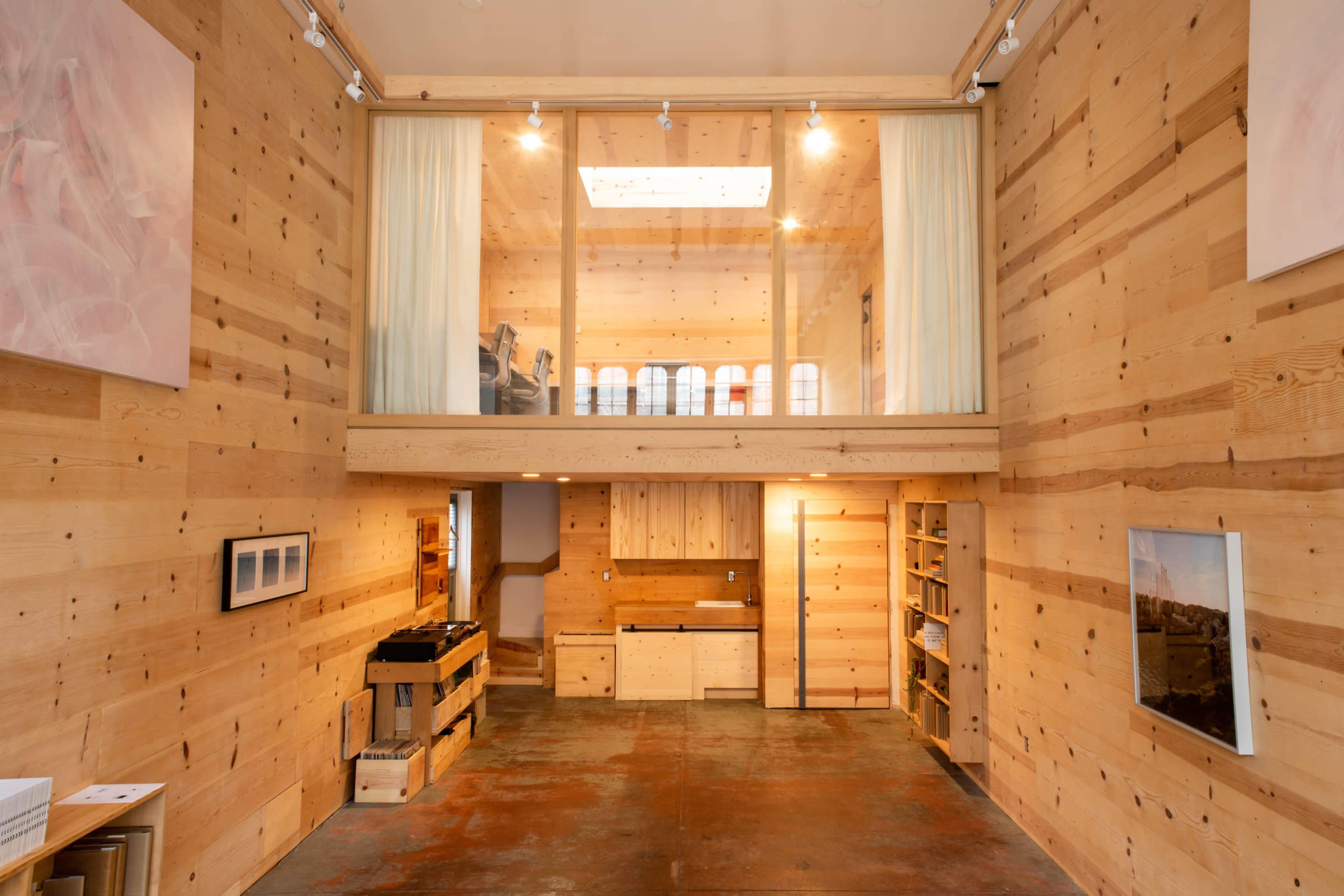 A spacious interior of a wooden room with a mezzanine level, featuring minimalist furniture, large windows, and polished concrete flooring.