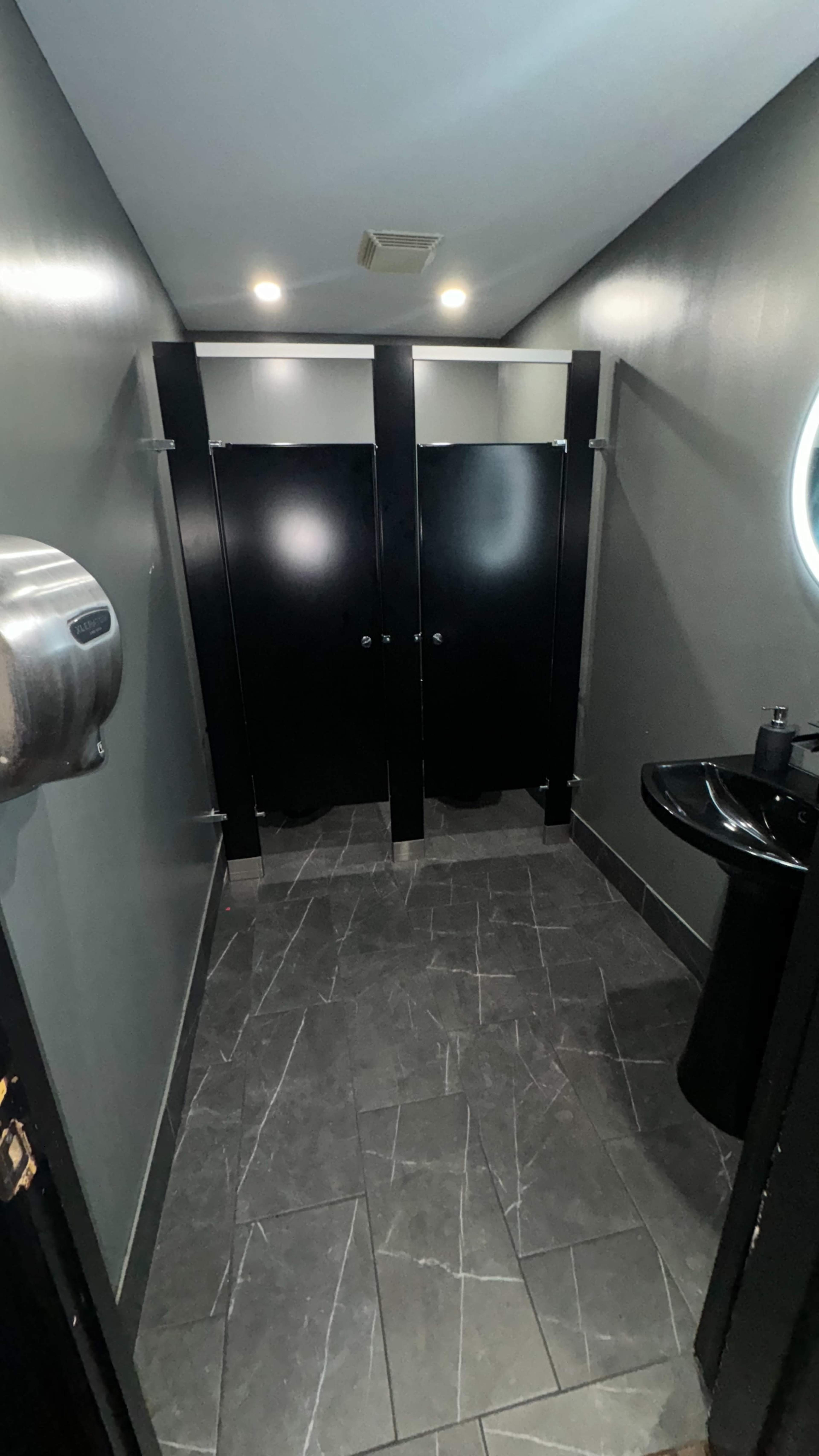 The image shows a narrow restroom with two black stall doors and a small sink with a mirror on the wall.