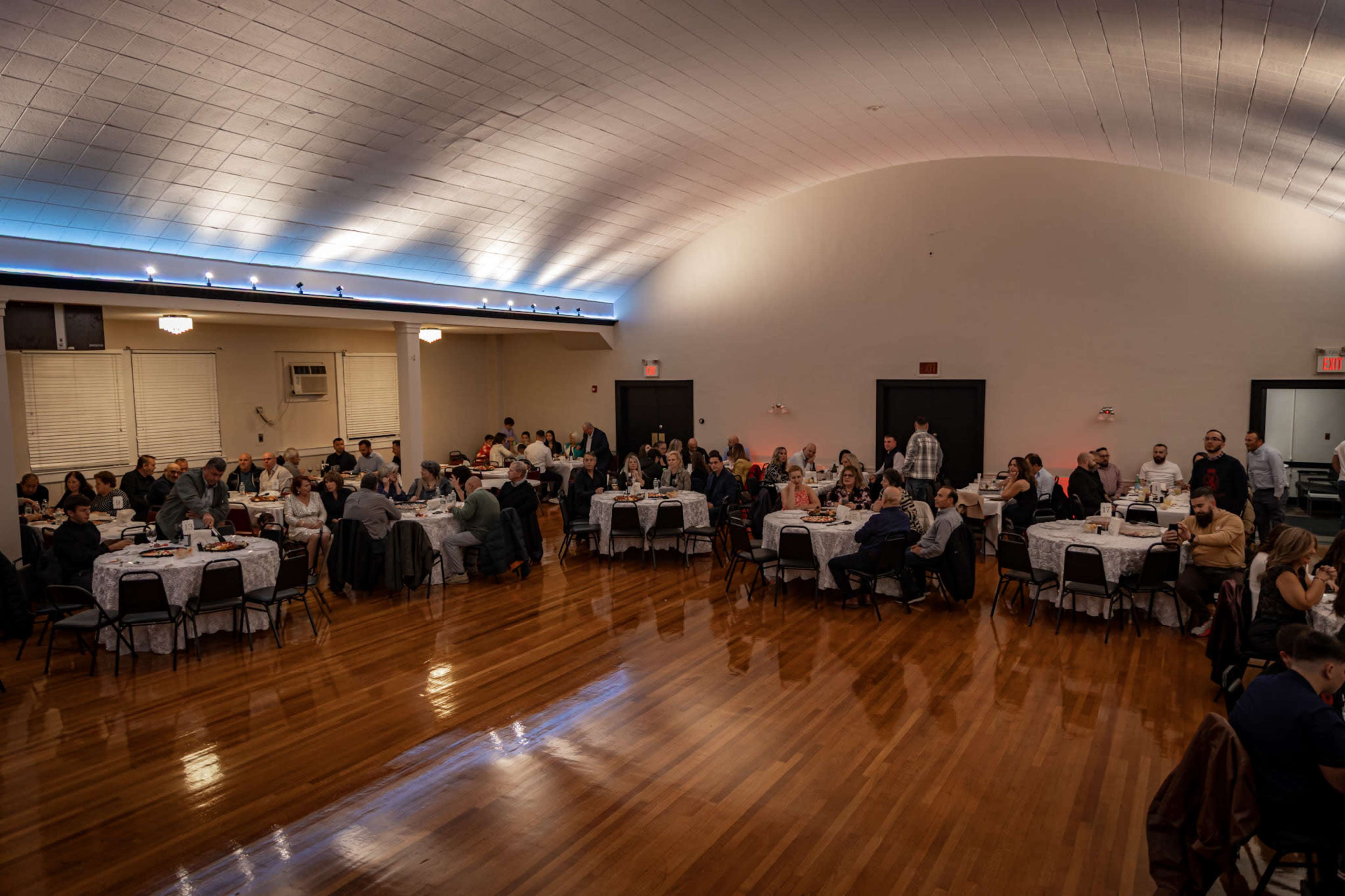 A large banquet hall is filled with tables set for dinner, with numerous guests seated and interacting across the space.