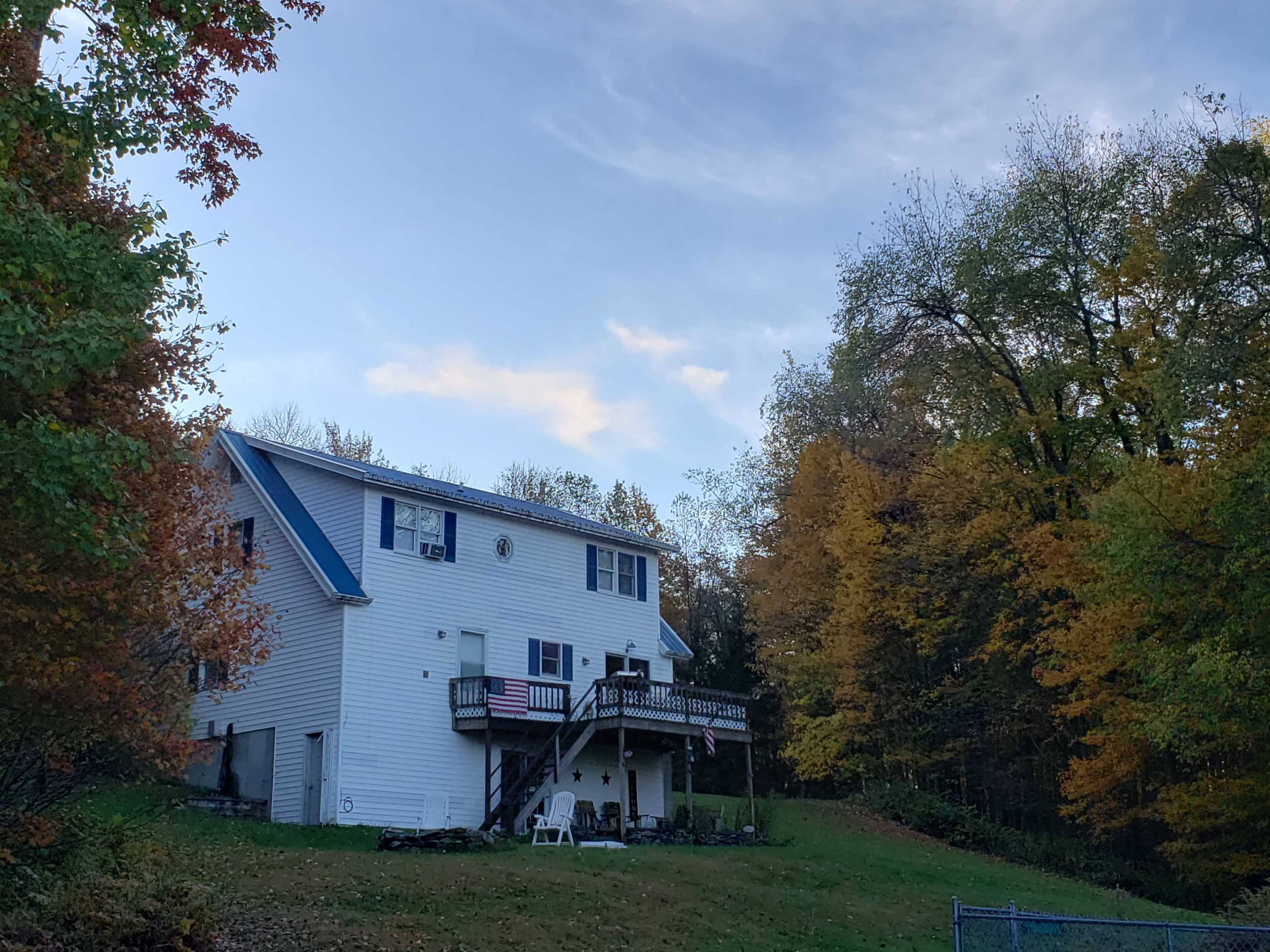 A two-story white house with blue accents is situated on a hillside surrounded by autumn-colored trees, featuring a deck with American flags.