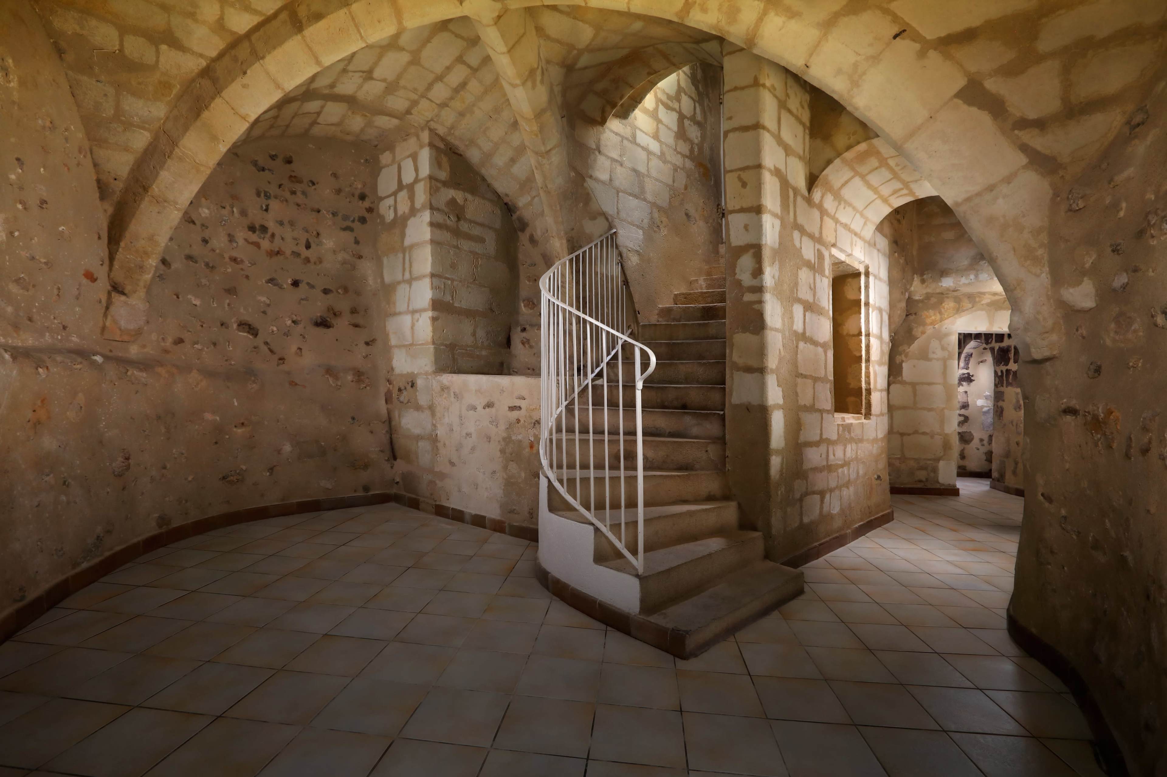The image shows a stone-walled interior with a curved staircase leading up, surrounded by arched ceilings and a tiled floor.