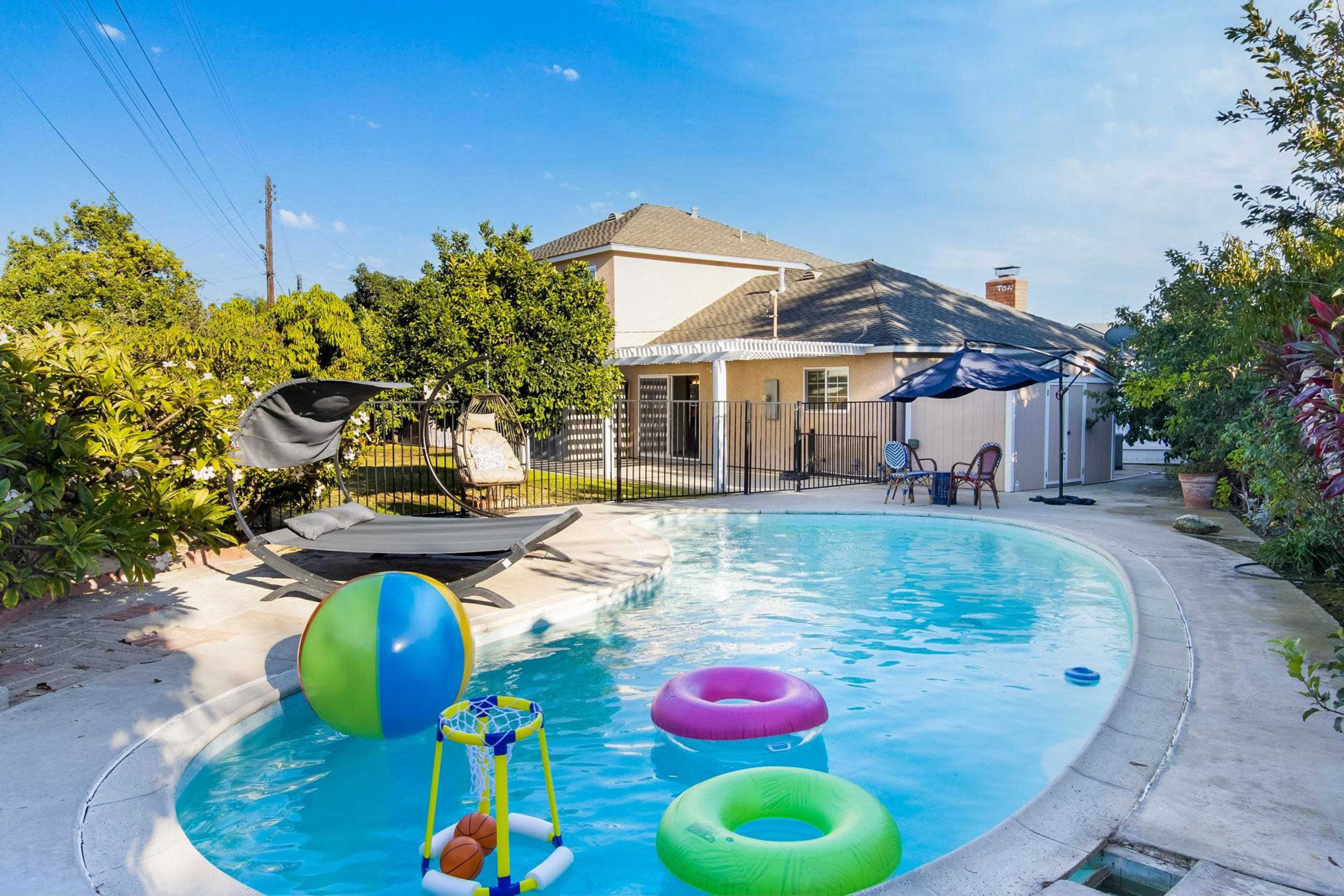 A backyard pool area with colorful inflatable toys and lounge chairs, surrounded by green trees and a fence.