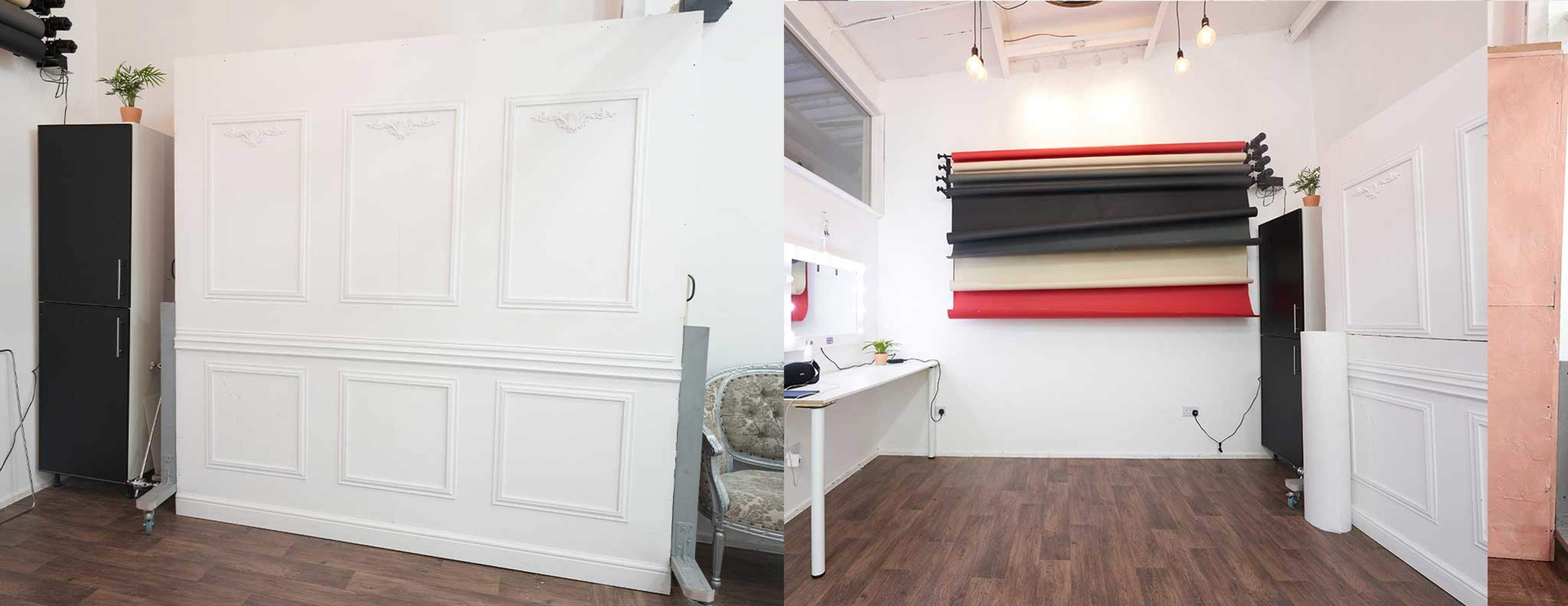 A bright studio space with a white wall featuring decorative paneling on one side and a table with various colored backdrop rolls mounted on the wall opposite.