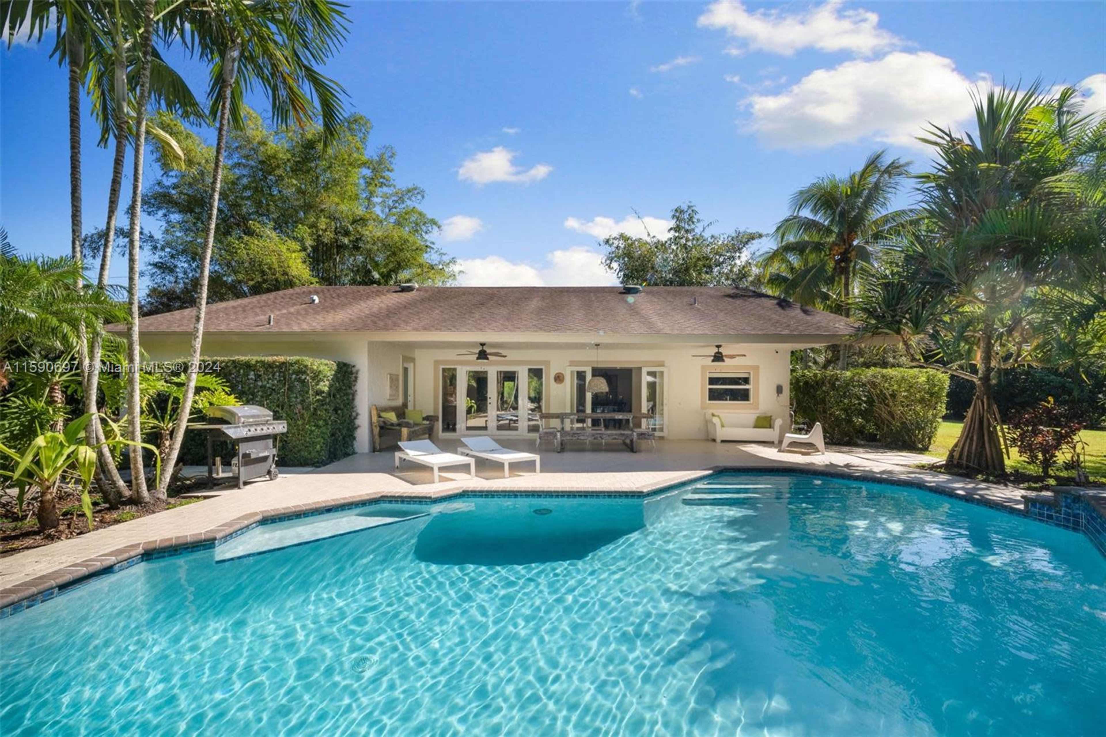 A modern backyard features a swimming pool, lounge chairs, and a covered patio with seating, surrounded by greenery and palm trees.