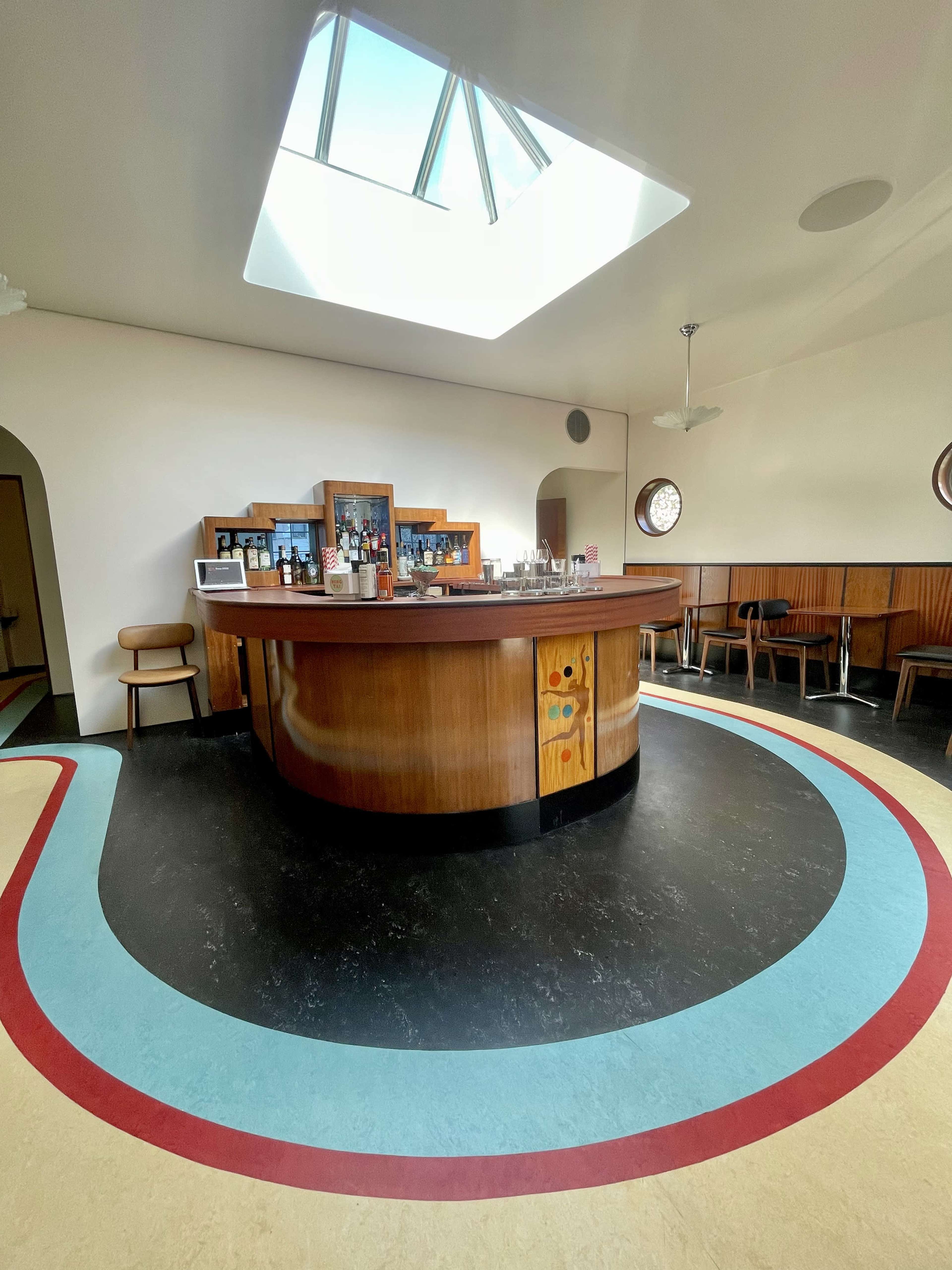 A modern bar with a wooden counter and circular patterned flooring beneath a skylight.