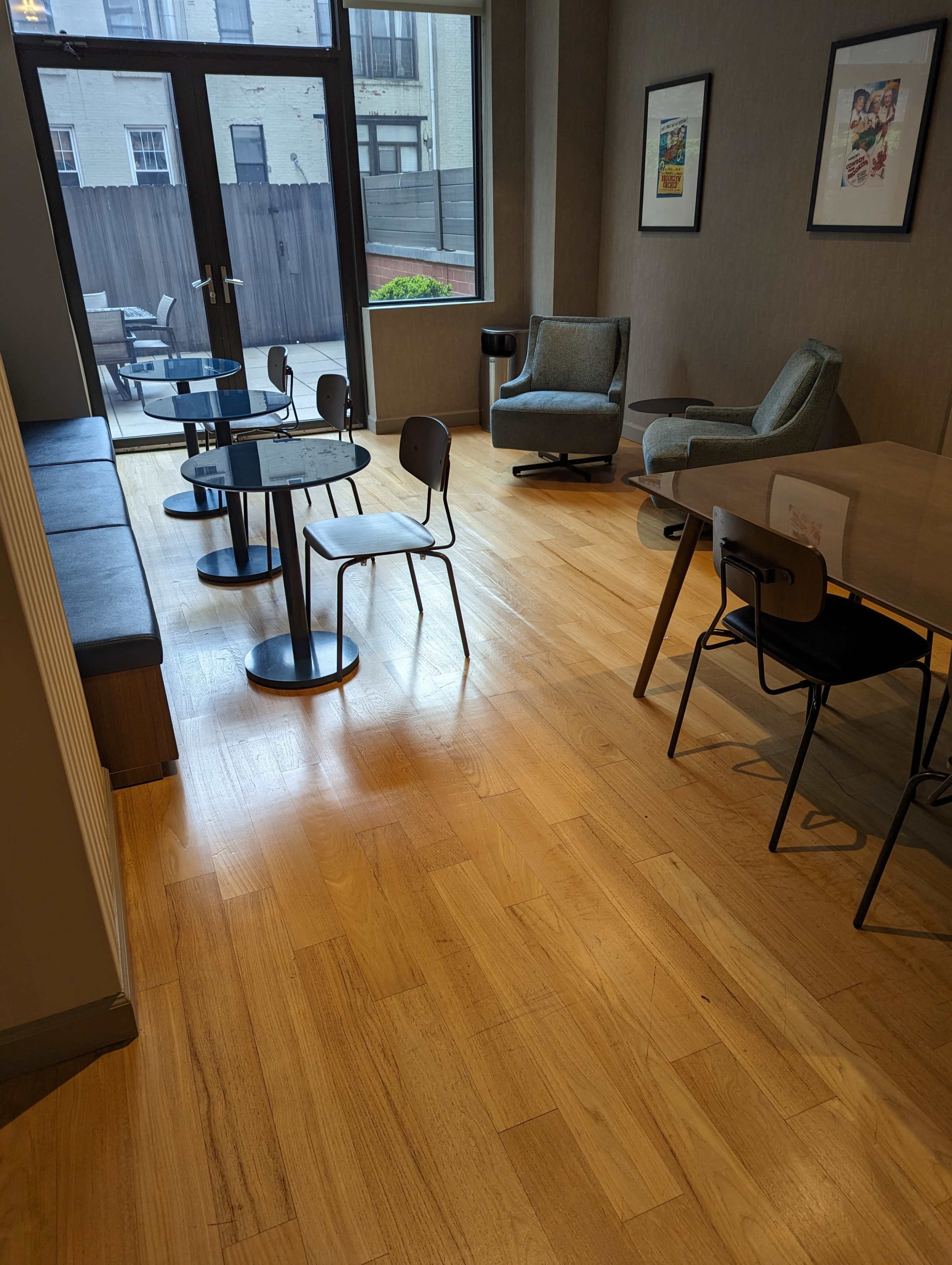 A modern indoor space featuring several small tables and chairs, a cozy armchair, and large windows allowing natural light to enter.