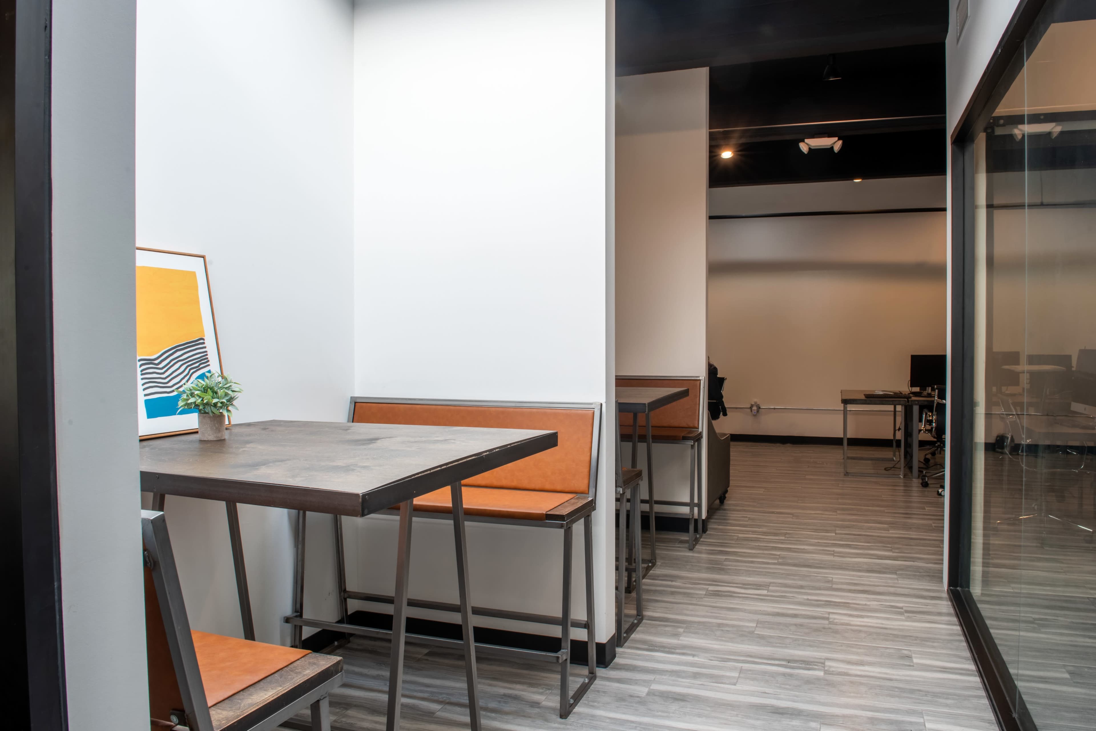 The image shows a modern office space featuring a small seating area with a table and benches, leading to a more open workspace in the background.