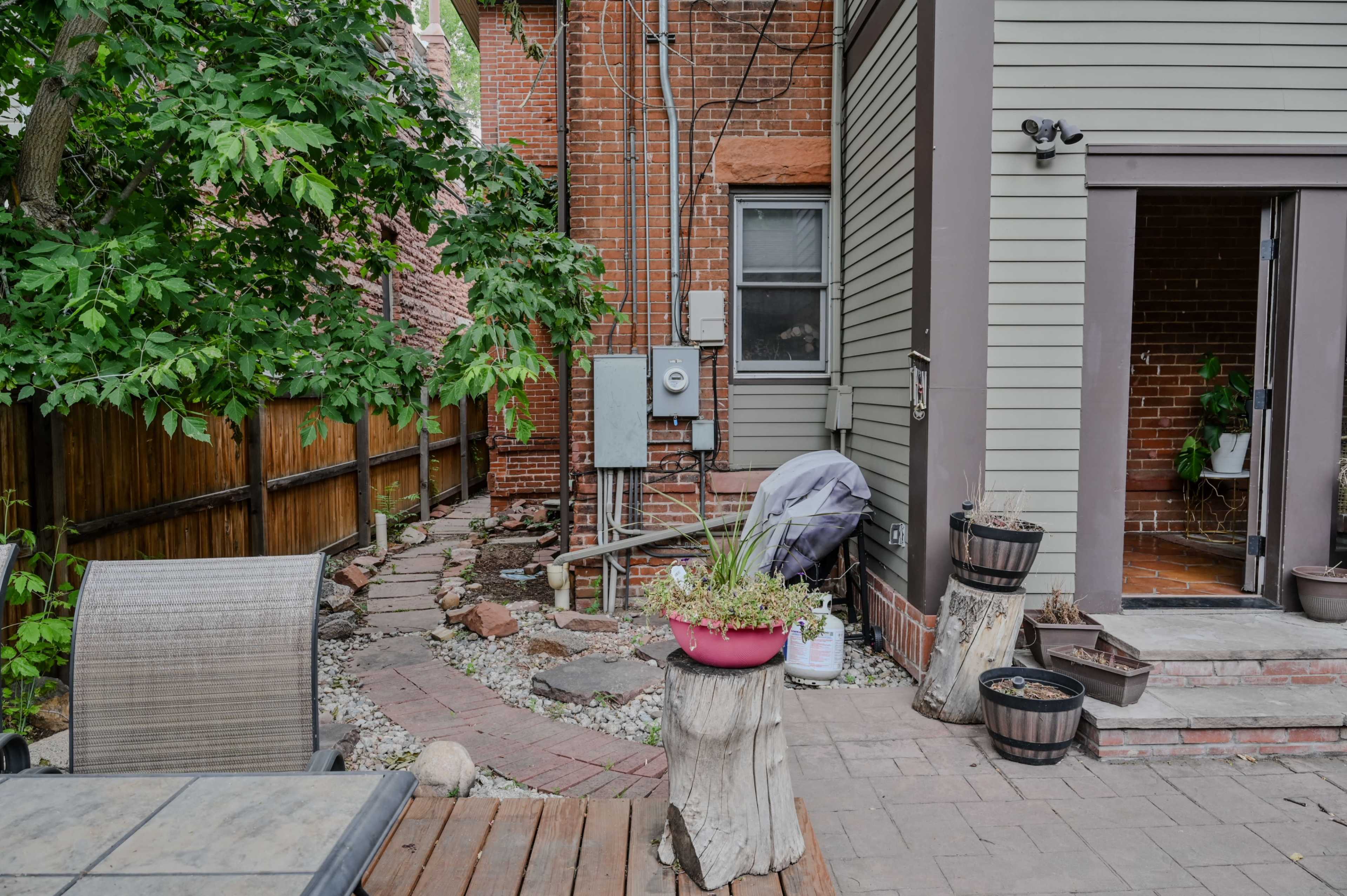 The image shows a backyard with a stone pathway, potted plants, and a utility area next to a wooden fence and brick buildings.