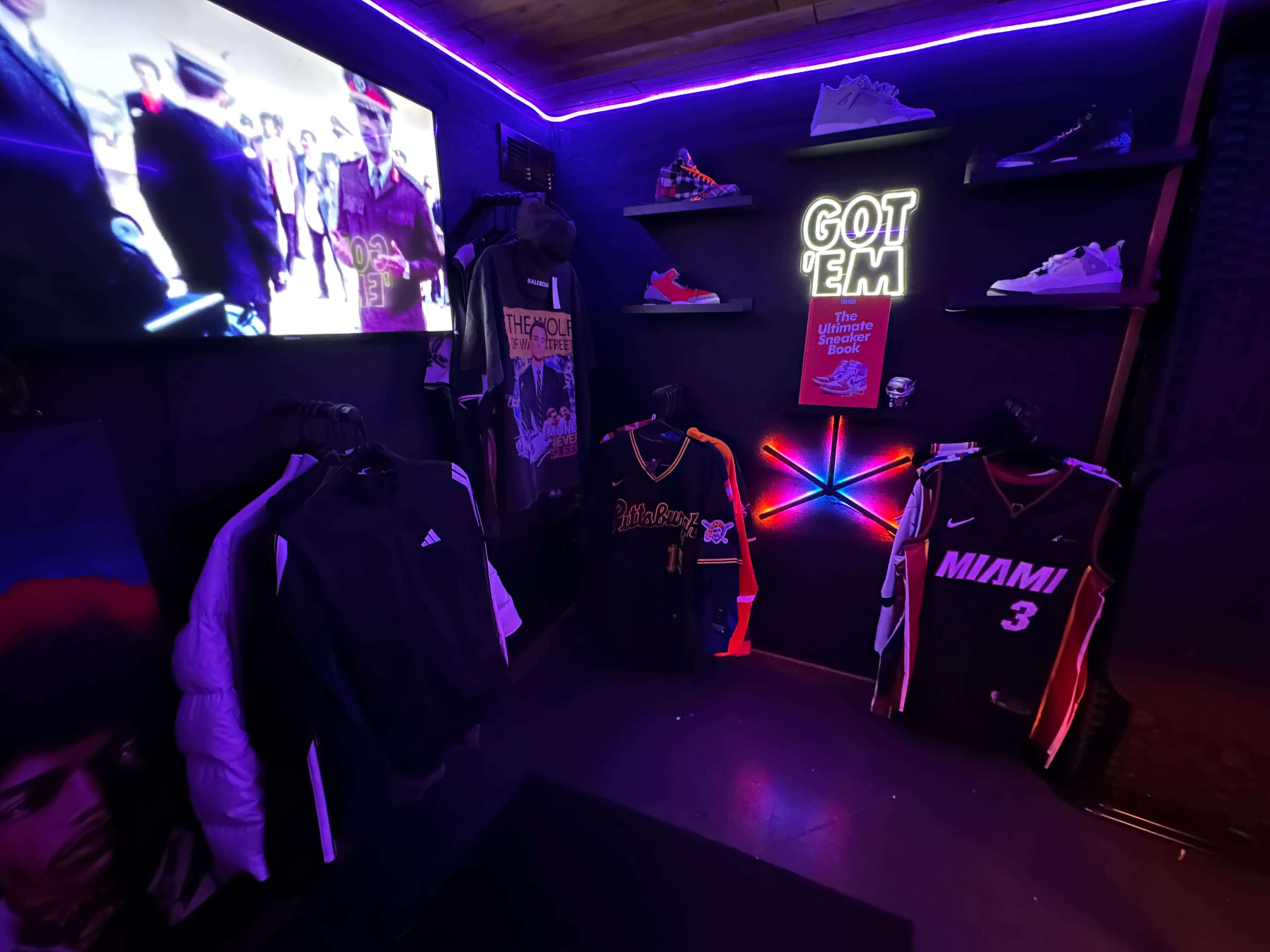 The image shows a brightly lit clothing display area featuring sports jerseys, jackets, and sneakers, with a television screen playing a video in the background.