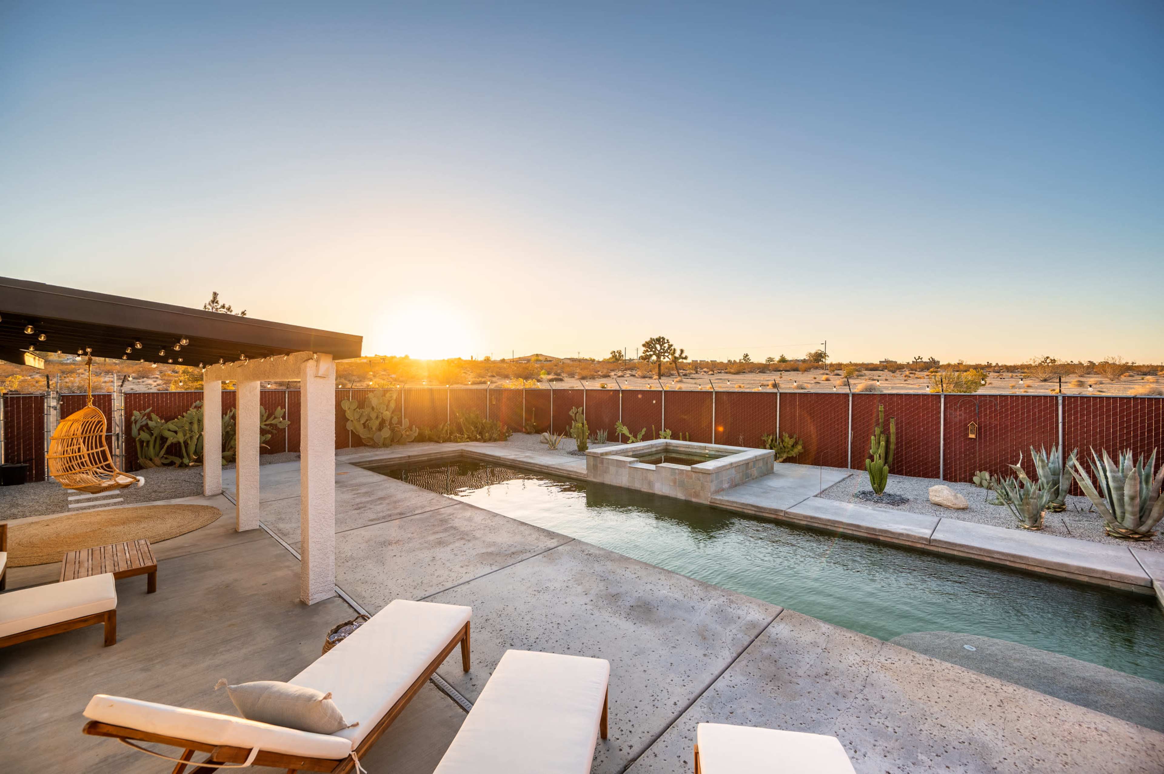 A swimming pool with lounge chairs sits in a desert landscape, surrounded by cacti and a view of the sunset.
