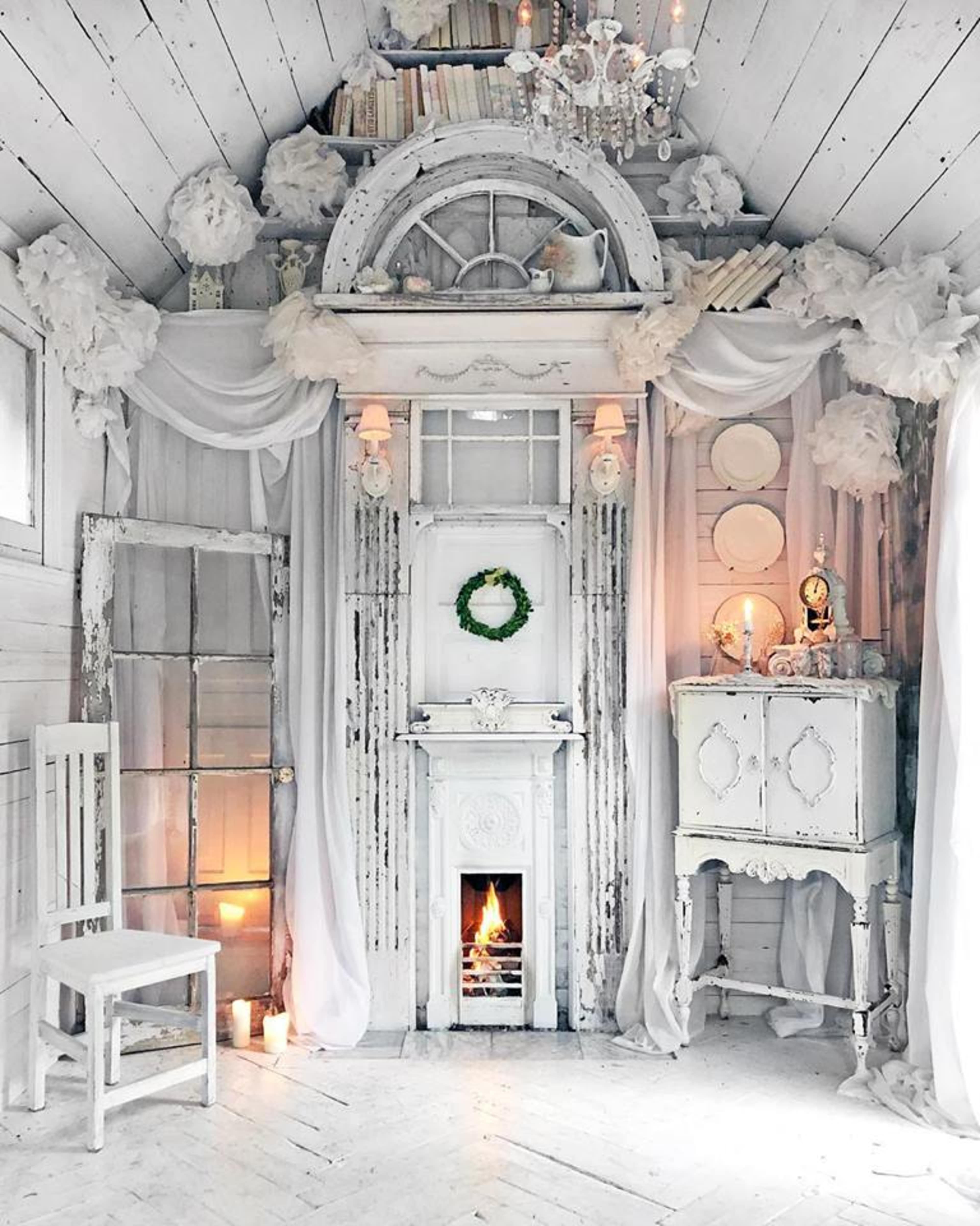 A white, elegantly decorated room featuring a vintage fireplace, a chandelier, and walls adorned with draped fabric and decorative accents.