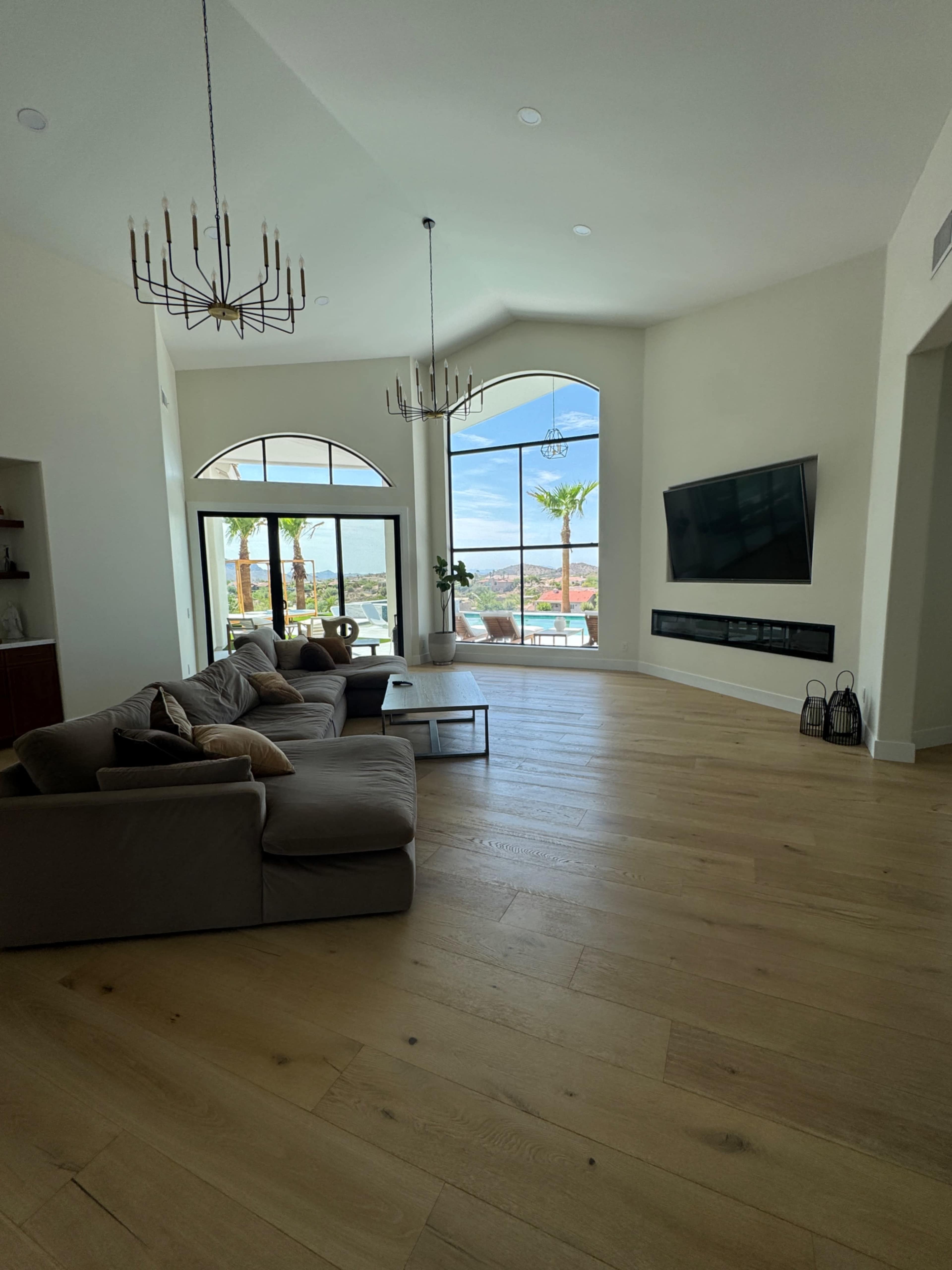 The spacious living room features a large sectional sofa, a mounted flat-screen TV, and floor-to-ceiling windows with views of palm trees and distant hills.