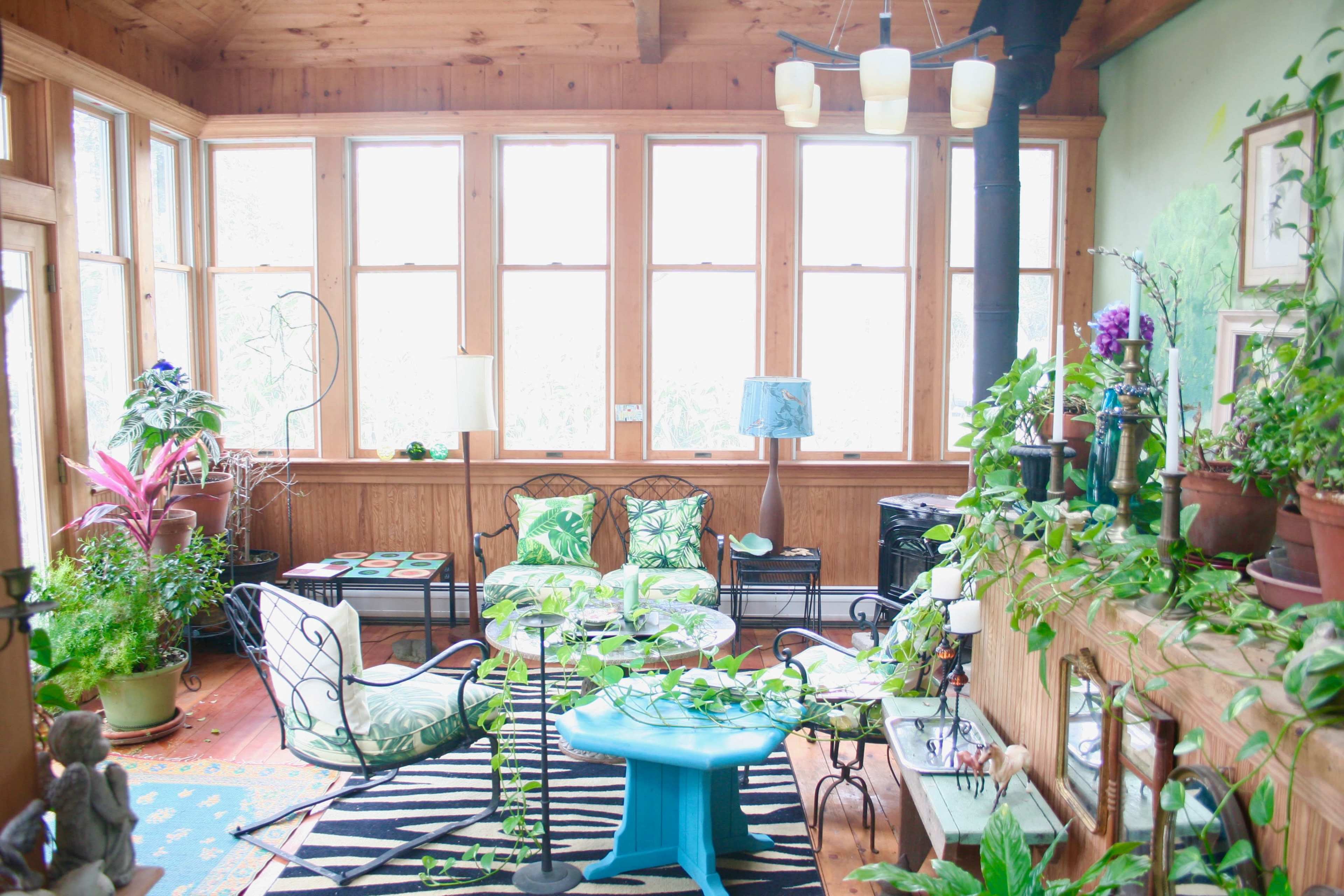 A bright sunroom filled with various indoor plants, wicker furniture, and large windows allowing natural light to illuminate the space.