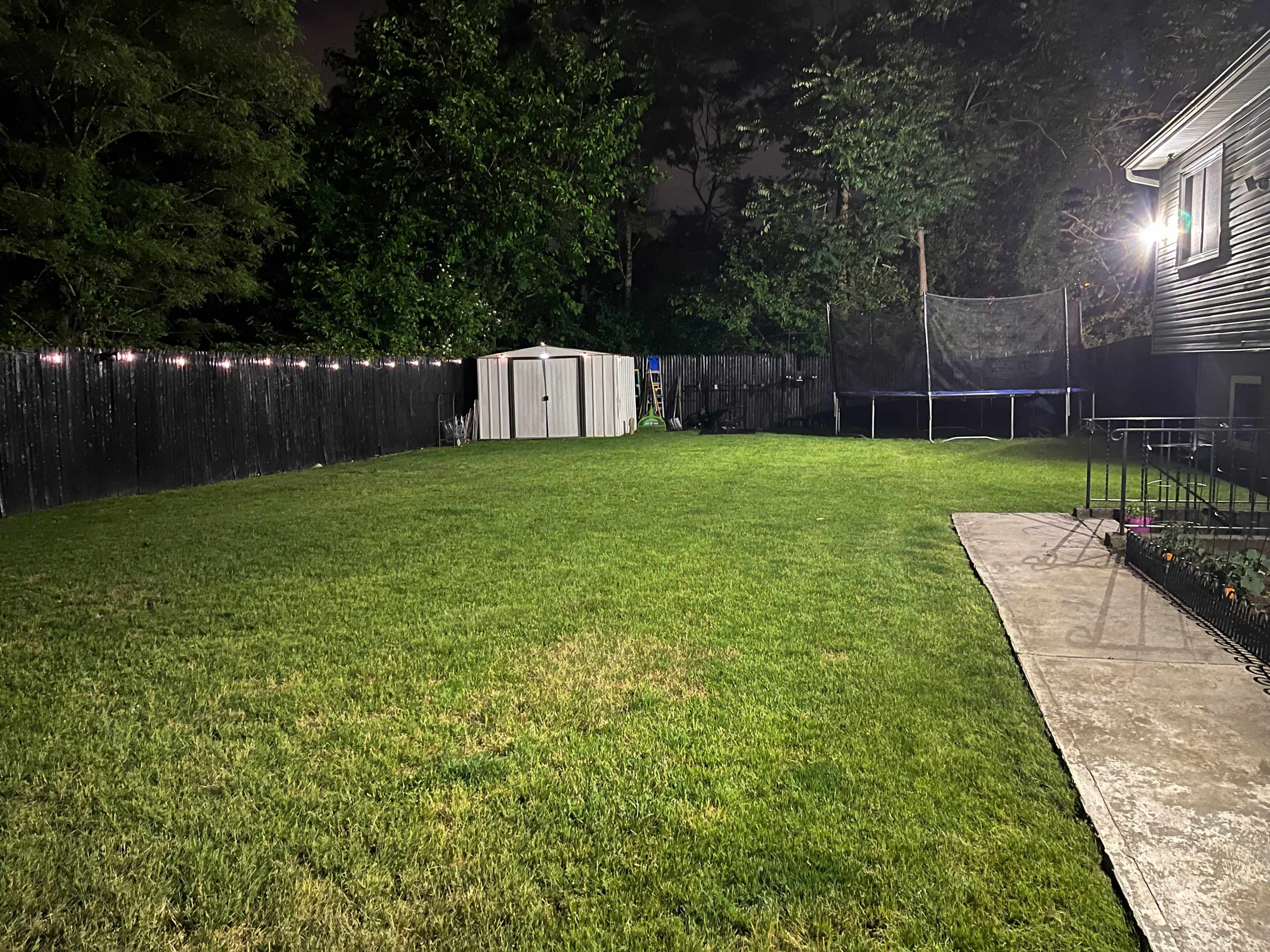 A large, well-mowed backyard at night contains a shed, a trampoline, and a pathway leading to a house.