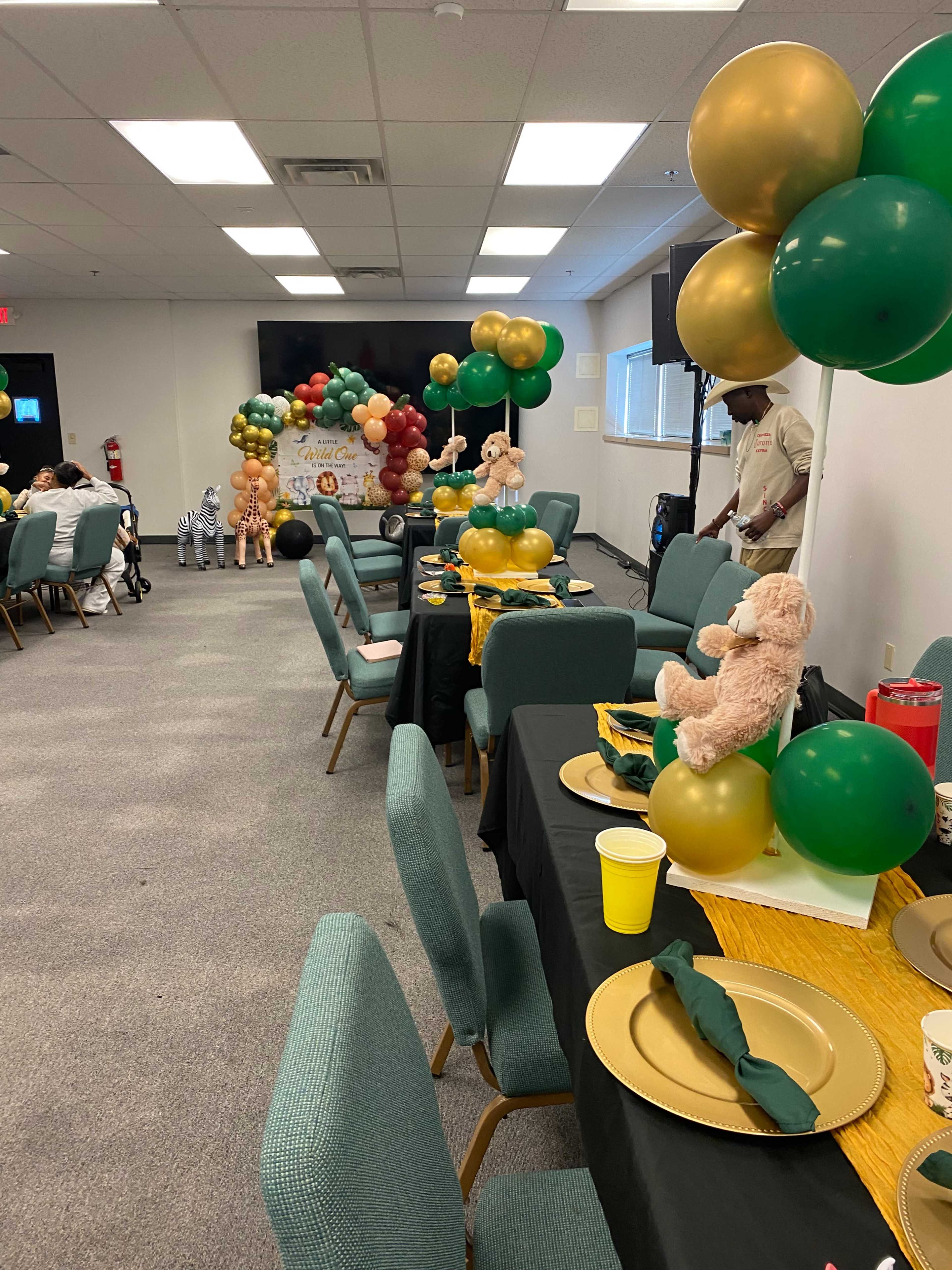 A decorated room features tables set with gold and green tableware and balloons, alongside plush toy animals for a celebration.
