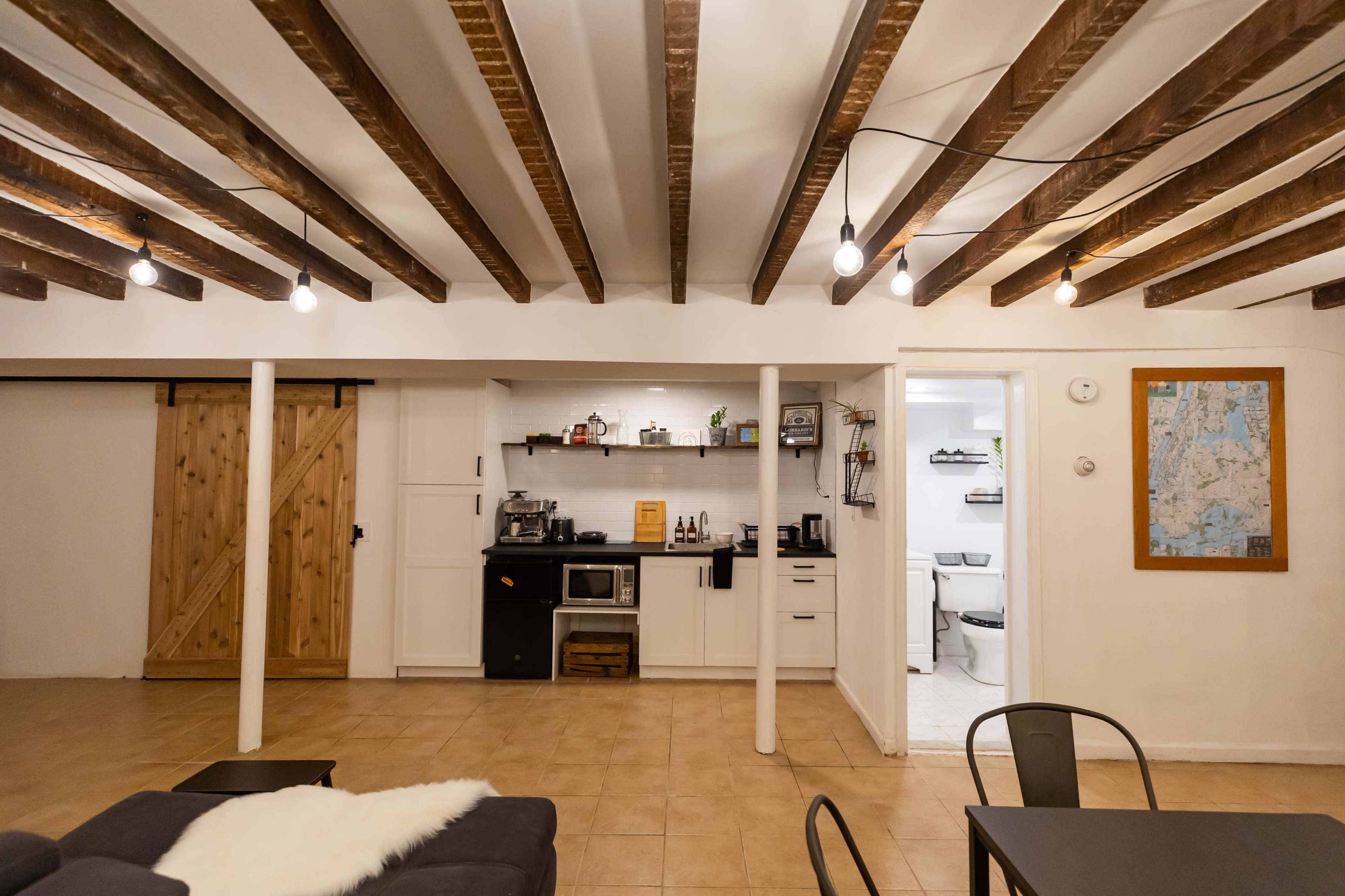A finished basement with exposed wooden beams, a kitchen area featuring white cabinets and a black stove, and a living space with a gray couch and light fixtures hanging from the ceiling.