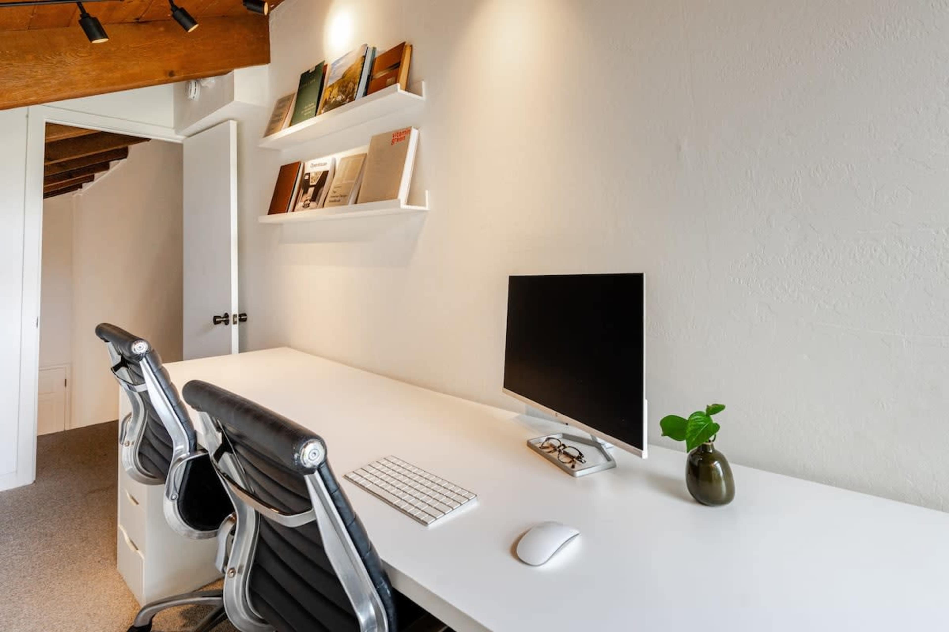 The image shows a minimalist office space featuring a long white desk with two office chairs, a computer monitor, a keyboard, and a small plant in a vase.