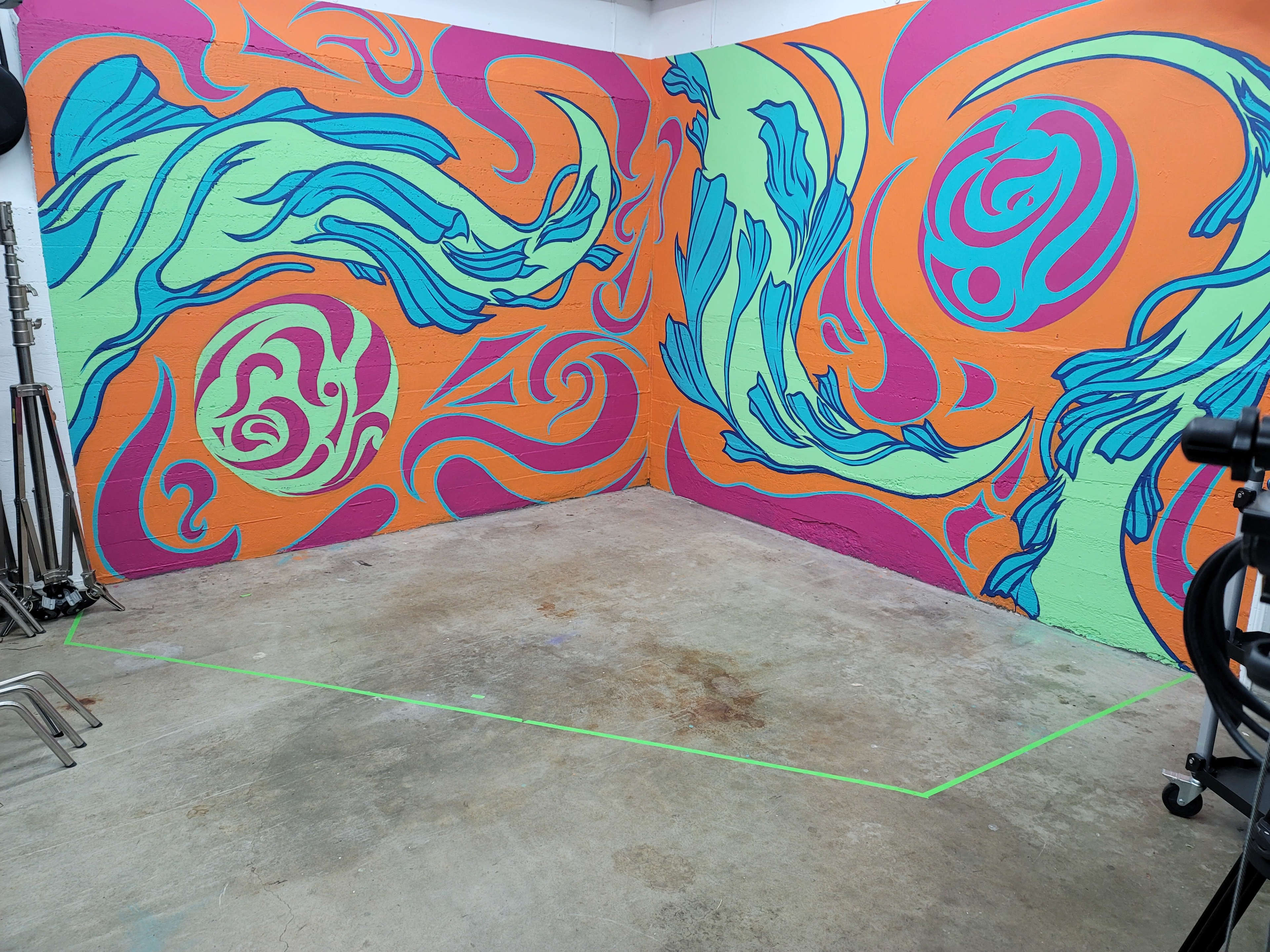 The image shows a room with brightly colored abstract murals on two walls and a concrete floor marked with green tape.