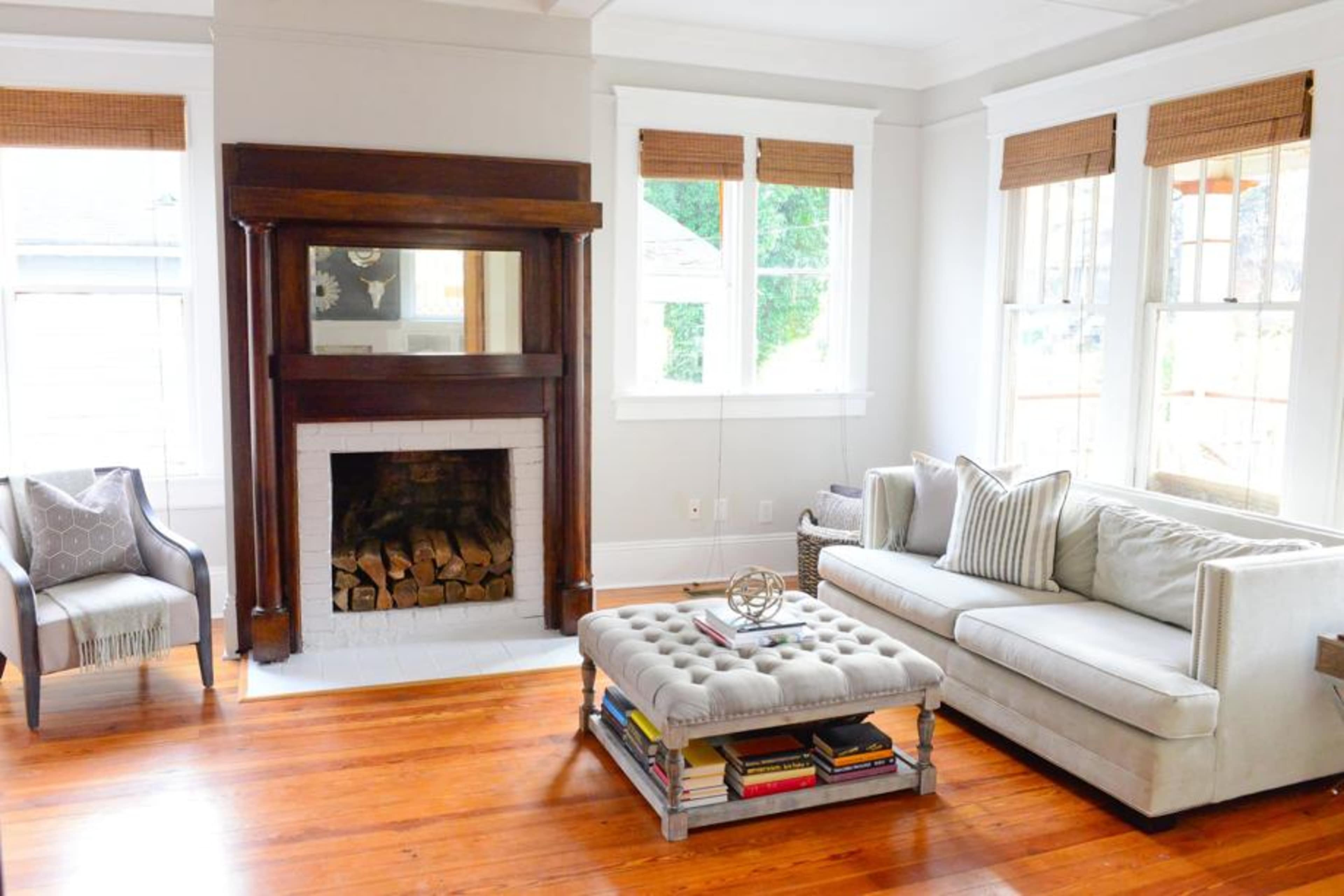 The room features a beige sofa and a tufted coffee table, with a wooden fireplace and large windows allowing natural light.