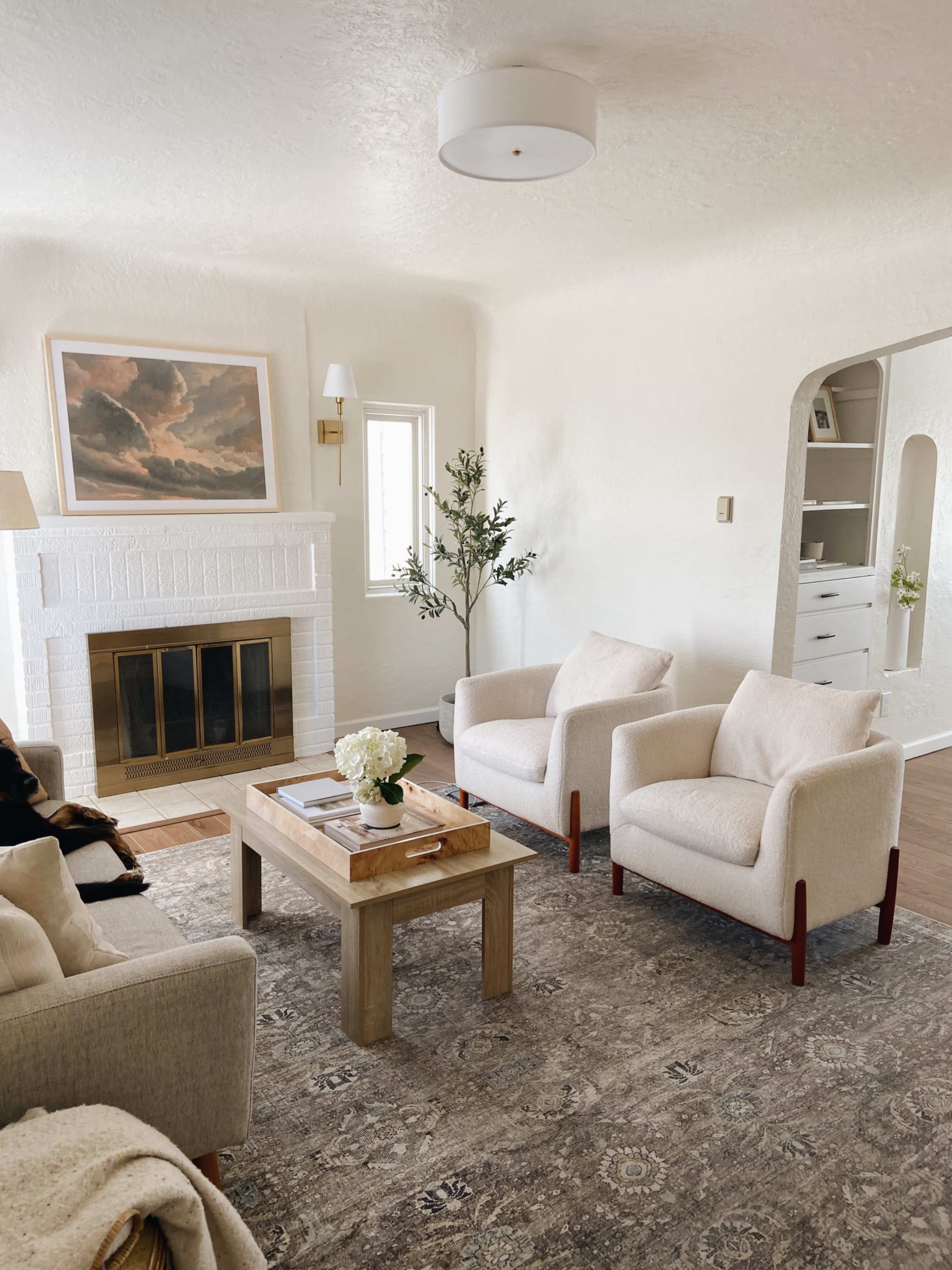 The living room features a white brick fireplace, two light-colored armchairs, a wooden coffee table, and a wall-mounted light, all set against a neutral backdrop.