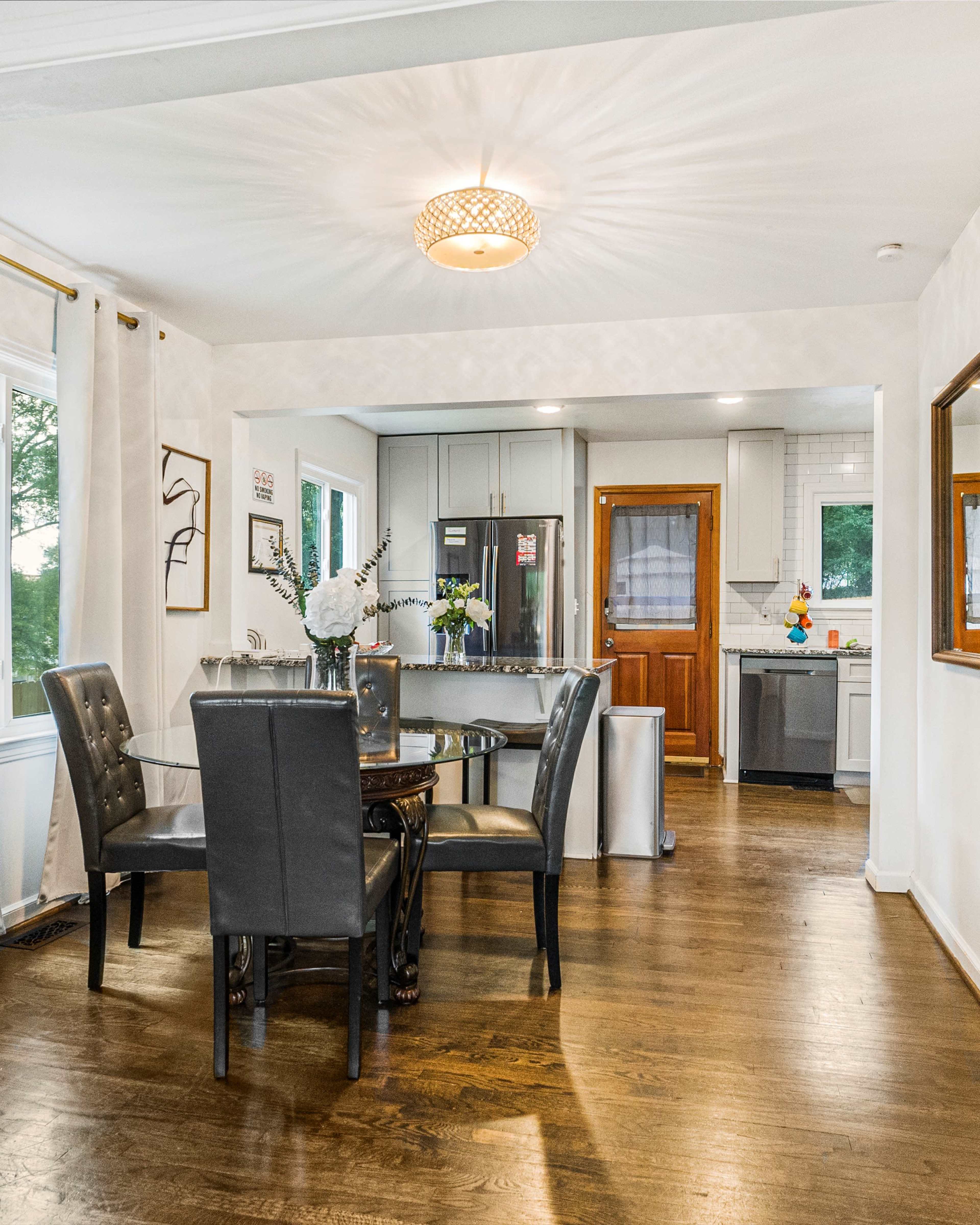 The image shows a modern dining area with a round glass table surrounded by black chairs, adjacent to a kitchen with stainless steel appliances.