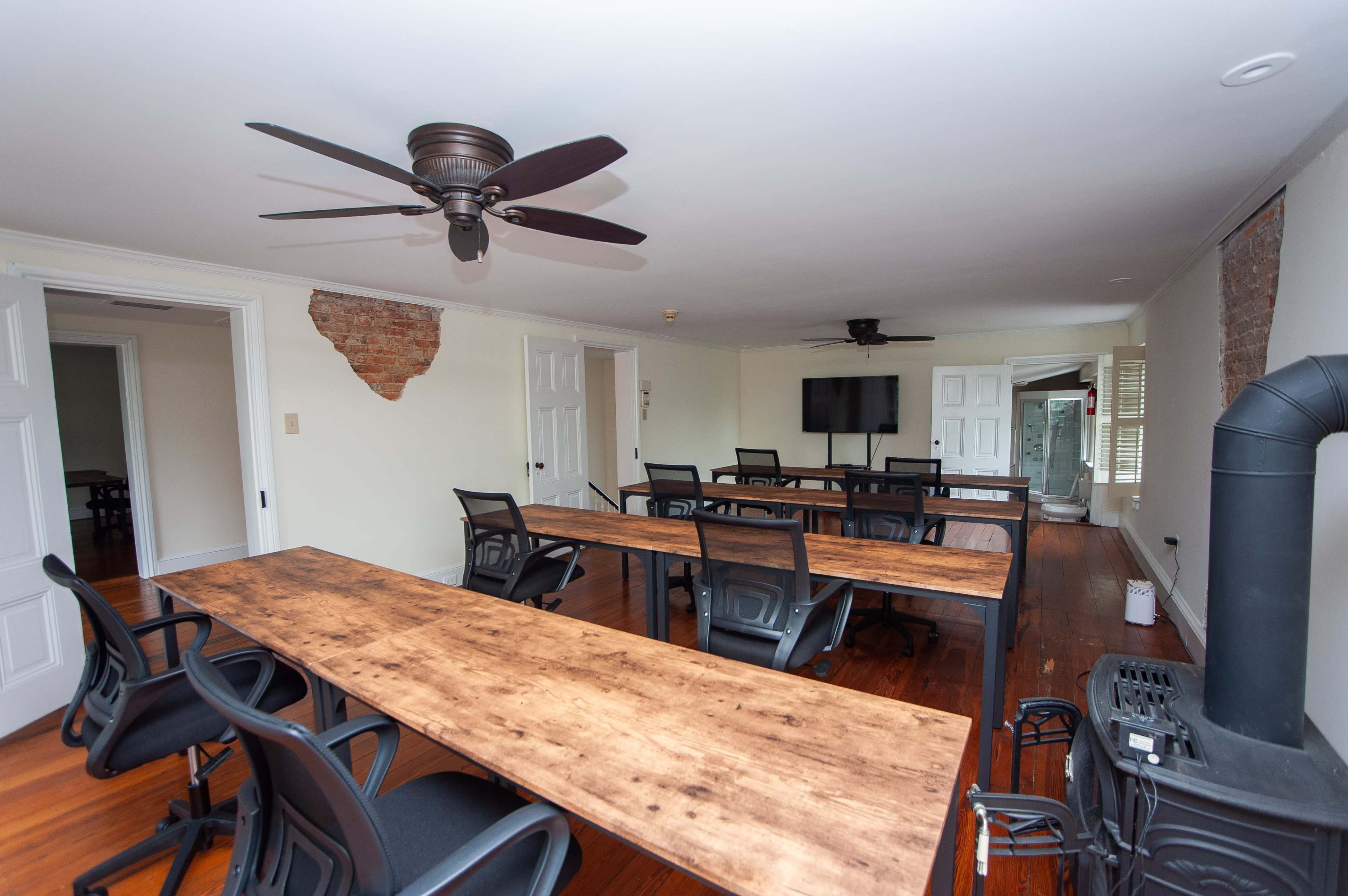 A workspace with multiple long tables and black chairs, featuring a ceiling fan and a television on the wall.