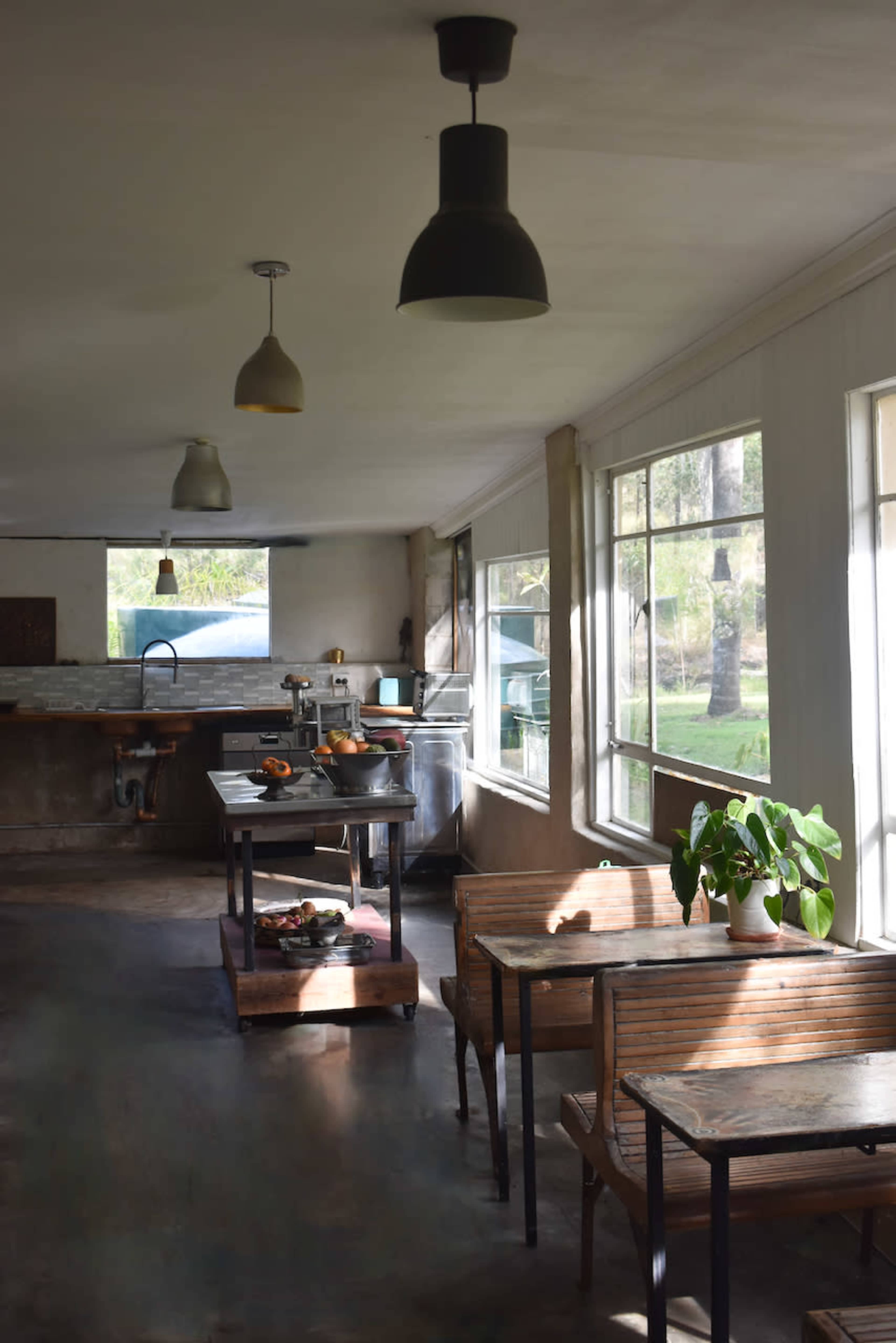 The image shows a spacious kitchen with a central table, wooden chairs, and large windows letting in natural light.