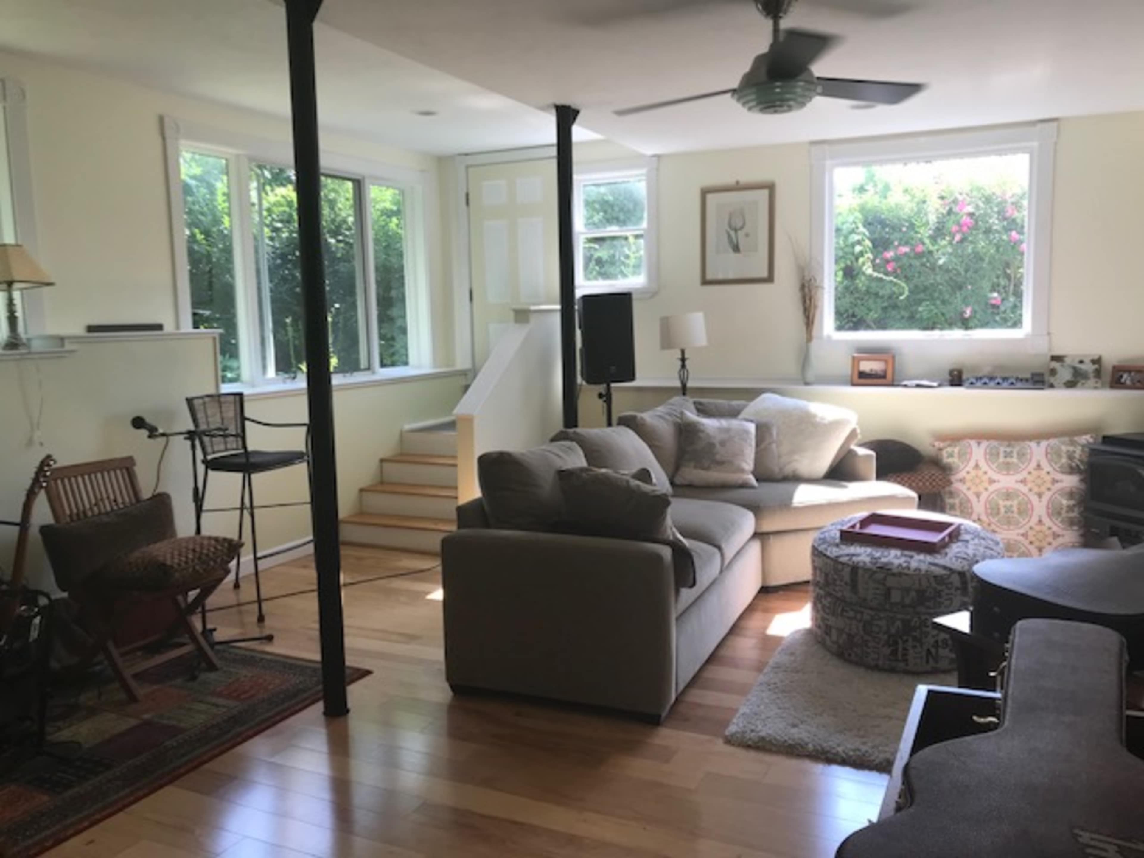 The image shows a living room with a sectional sofa, wooden floors, a coffee table, and large windows that let in natural light.