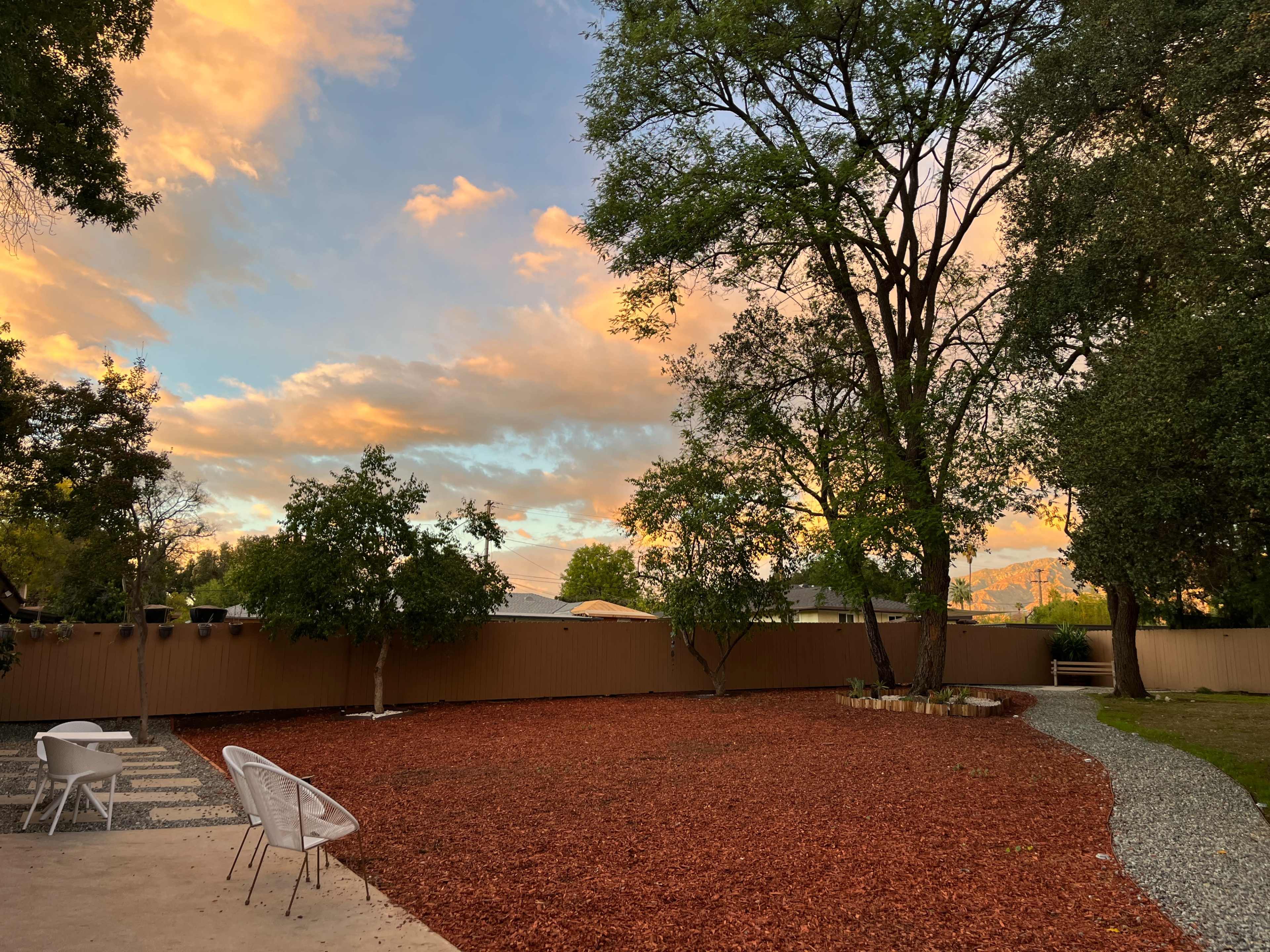 A backyard features a bed of red mulch surrounded by trees and a gravel path, under a colorful sunset sky.