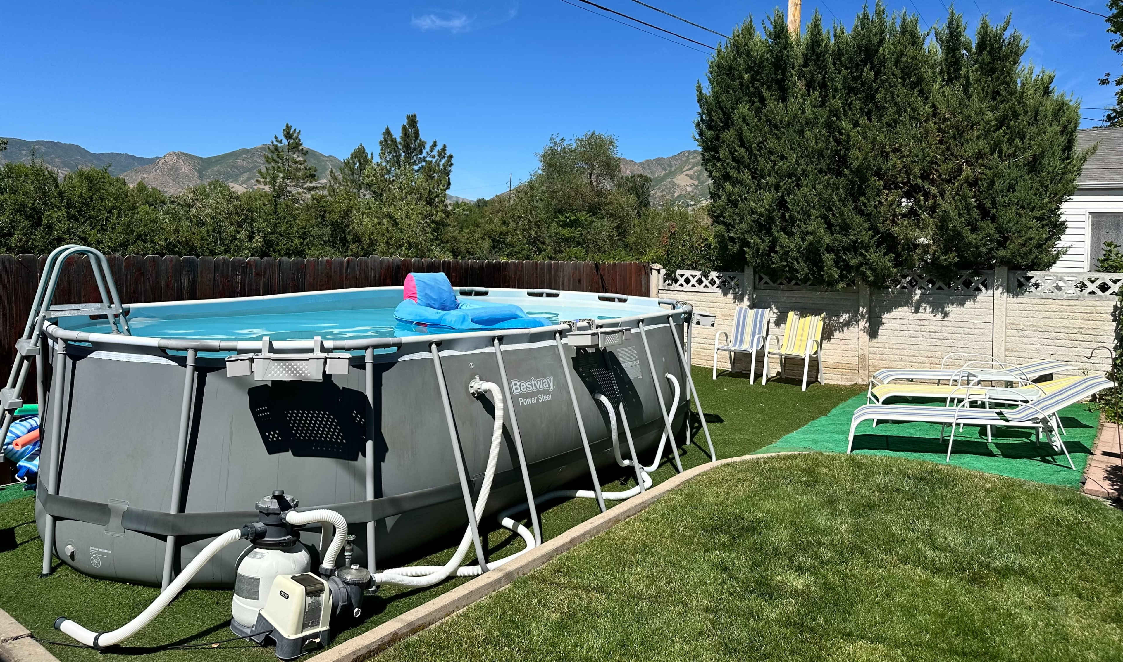 A round above-ground pool is set in a yard with green grass, surrounded by lounge chairs and shrubs under a clear blue sky.