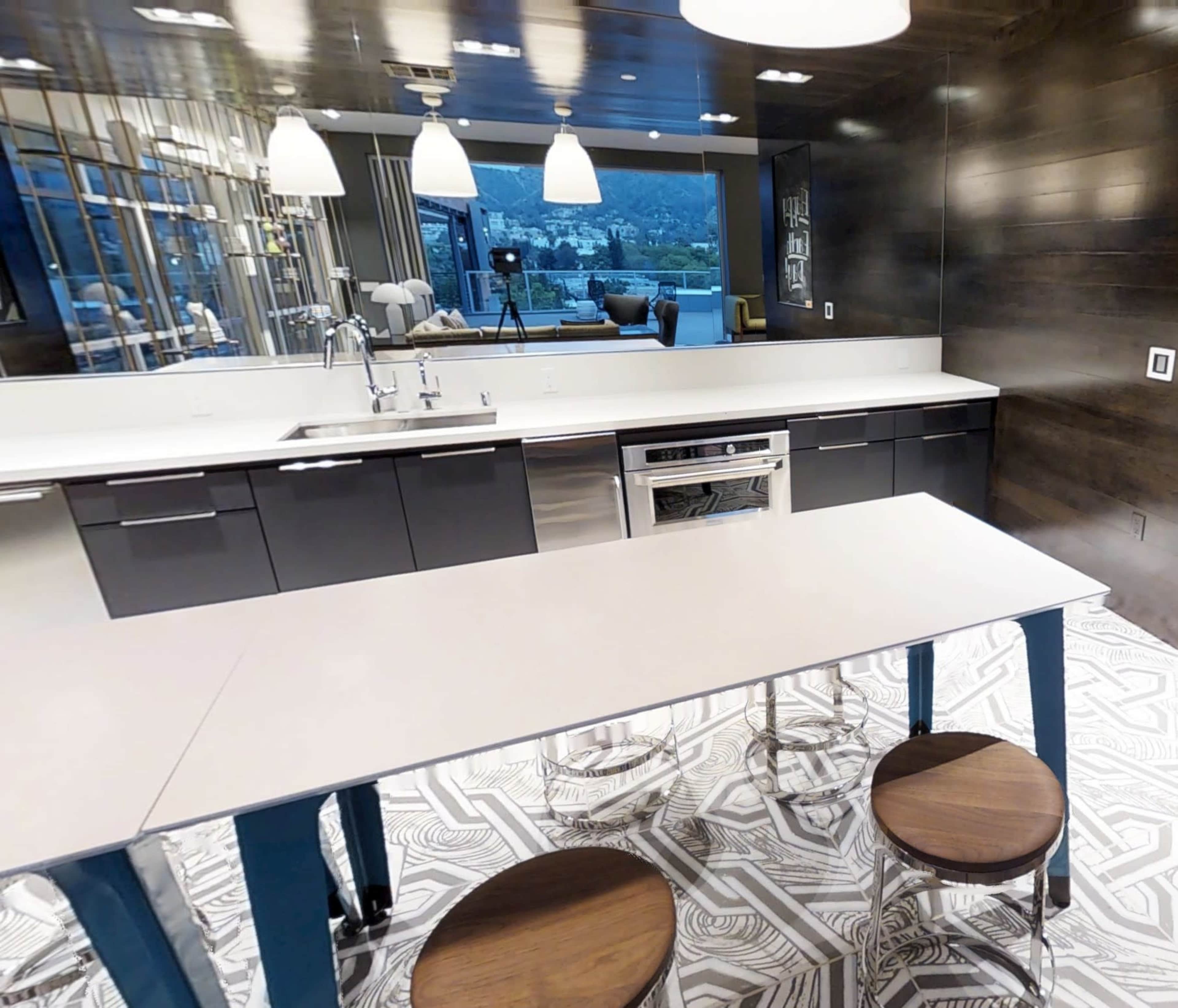 A modern kitchen features sleek black cabinets, a white countertop, and round wooden stools, with large windows overlooking a scenic view.