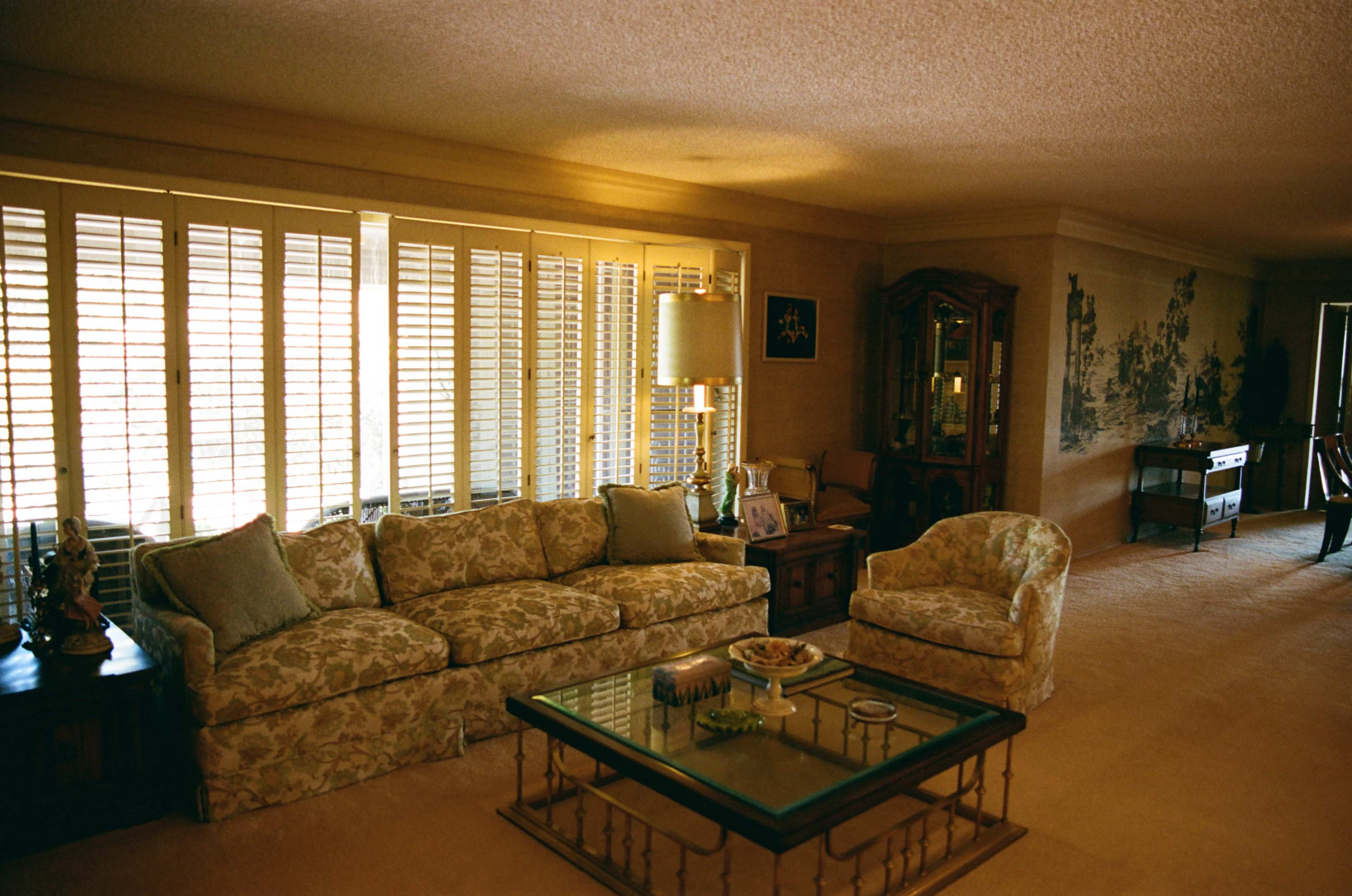 The image shows a living room furnished with a floral-patterned sofa, an armchair, and a glass coffee table, with large windows featuring wooden shutters.