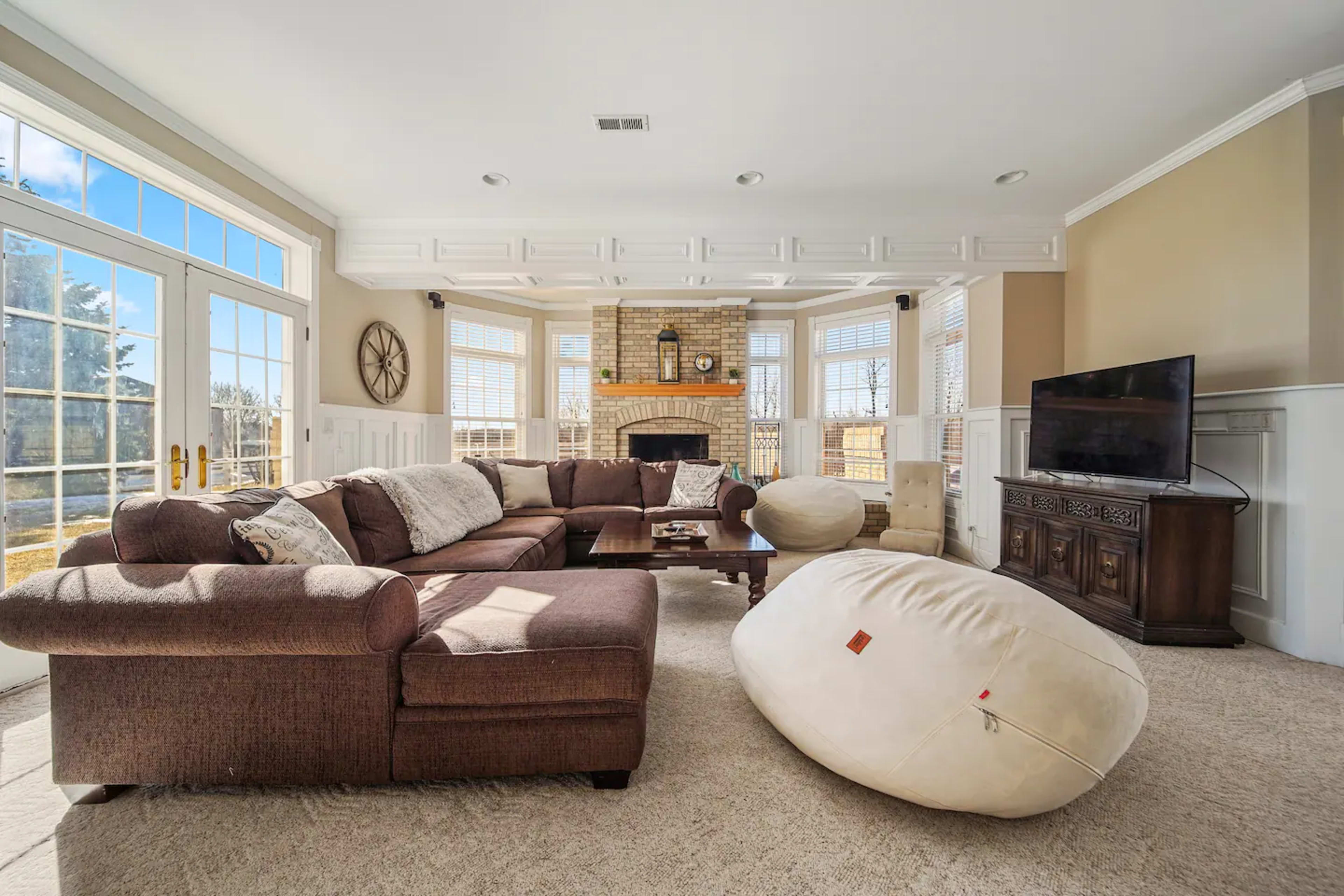 A spacious living room features a large sectional sofa, a coffee table, a flat-screen TV, and bean bag chairs, all set against a backdrop of large windows and a brick fireplace.