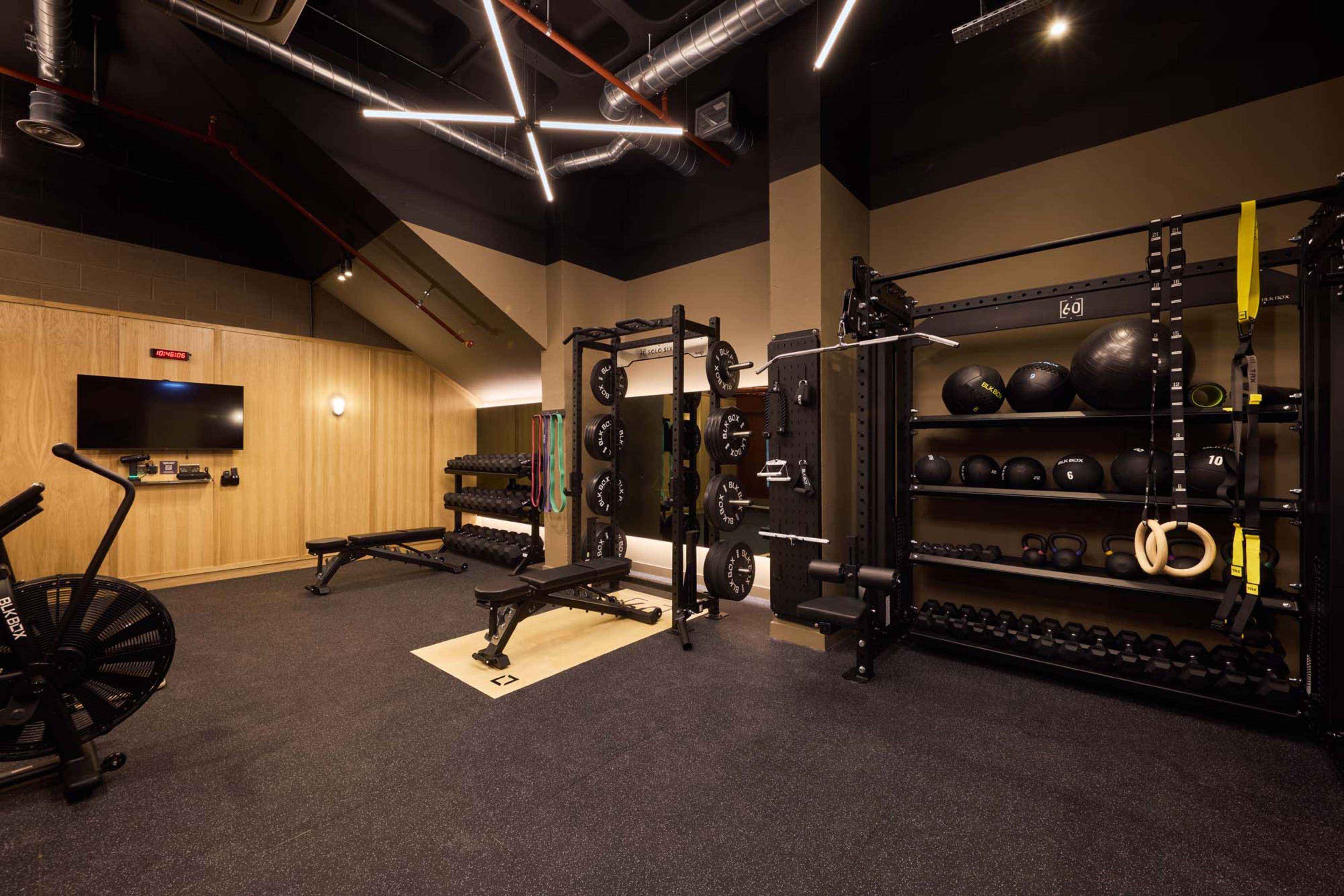 The image shows a modern fitness room equipped with various exercise machines, free weights, and a rack of medicine balls.