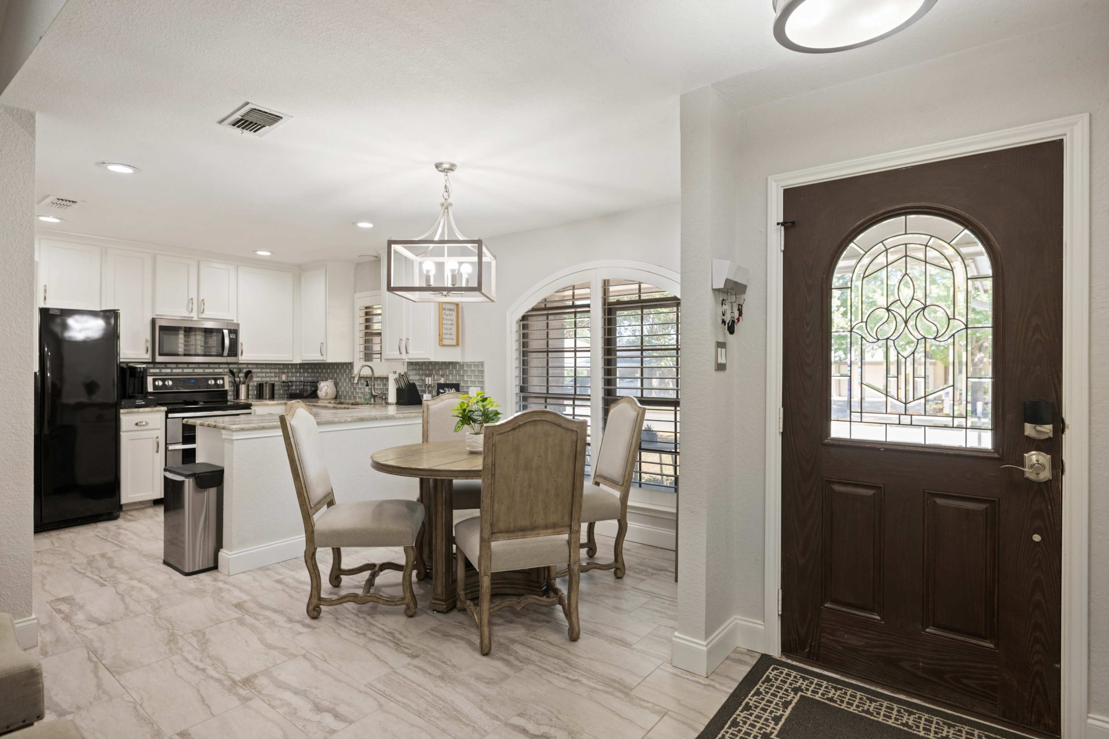A modern kitchen with a round dining table, four chairs, and a front door with decorative glass, all illuminated by natural light.