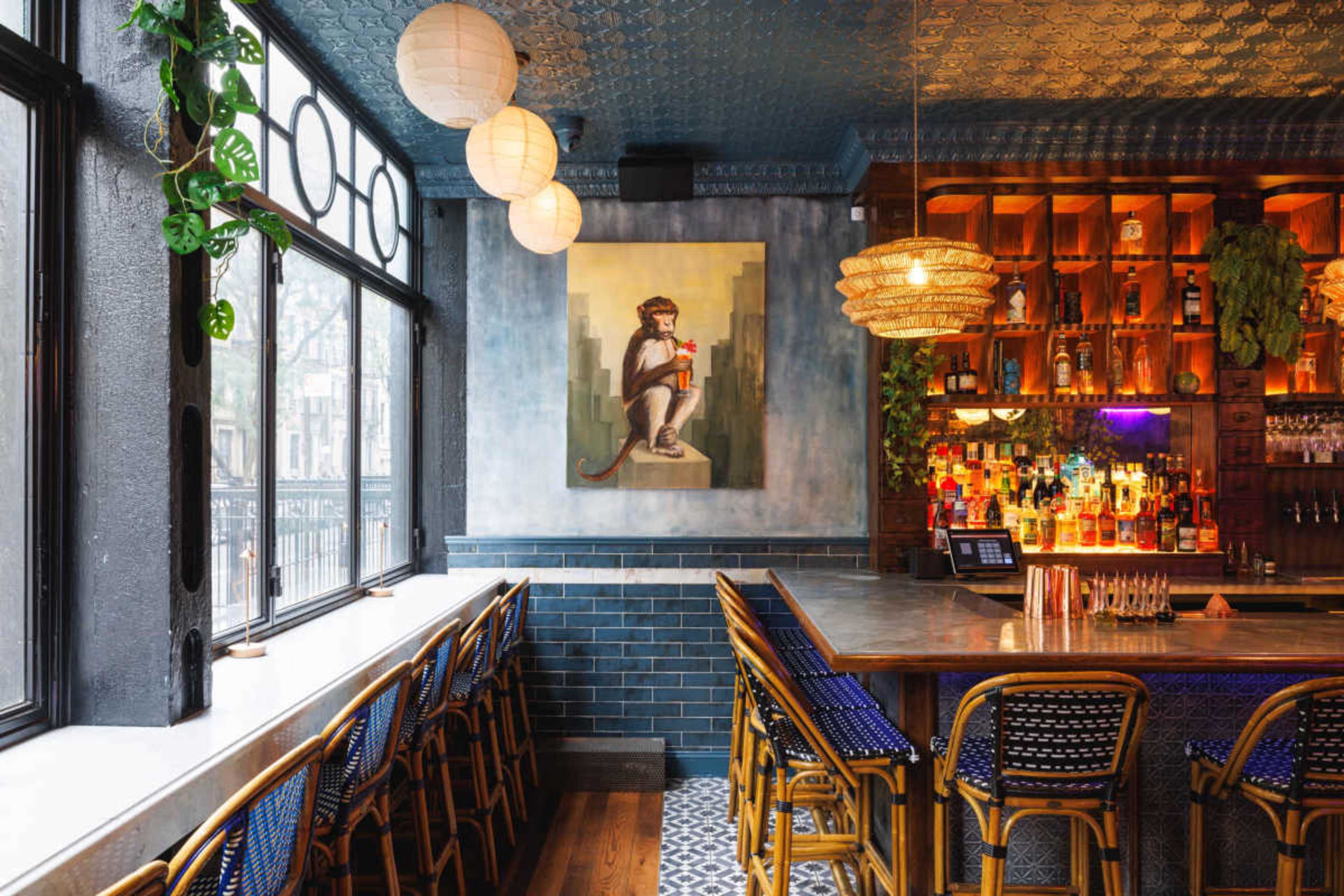 The image shows a stylish bar interior with a large painting of a monkey on the wall, a long counter with stools, and a wide selection of illuminated bottles behind the bar.