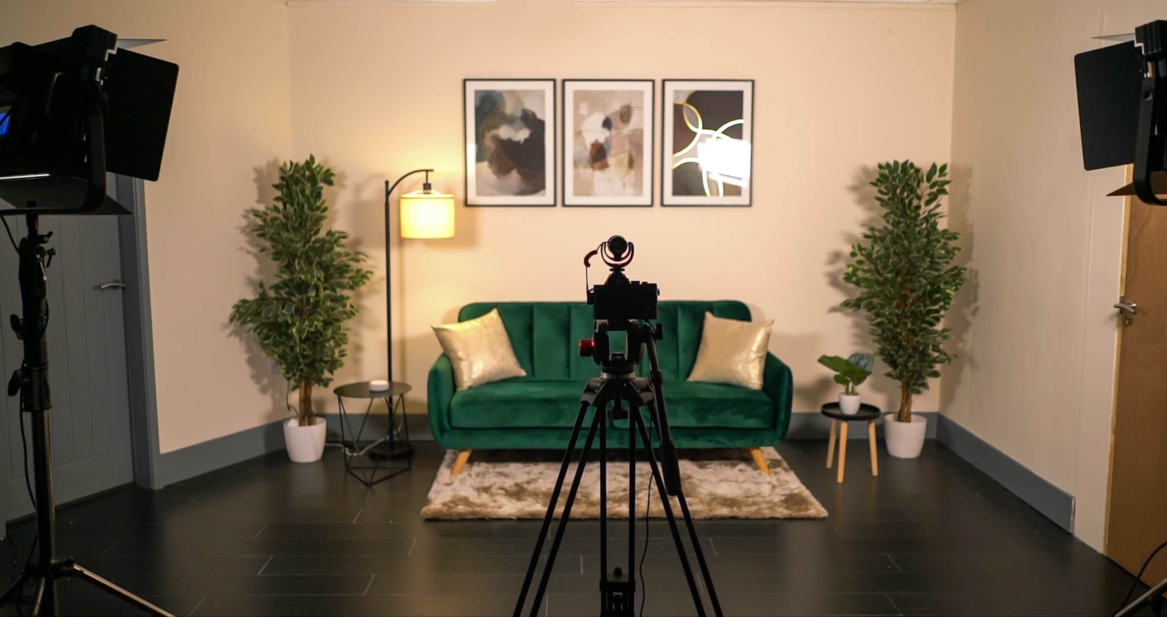 A neatly arranged studio setup featuring a green sofa, two potted plants, a lamp, and a camera mounted on a tripod facing a decorated wall.