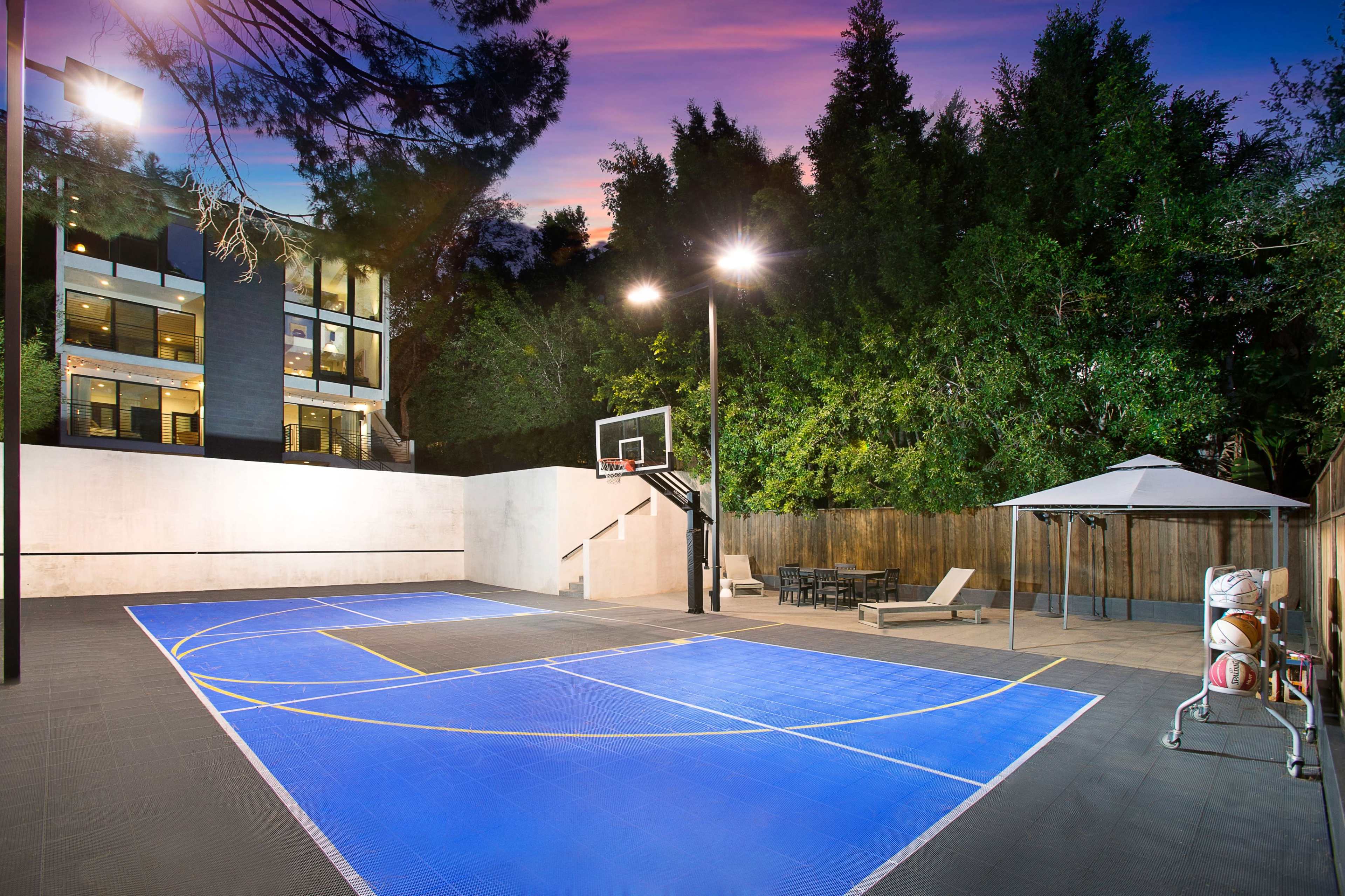 A basketball court with a blue surface, surrounded by trees and a seating area with a gazebo, against the backdrop of a modern building.