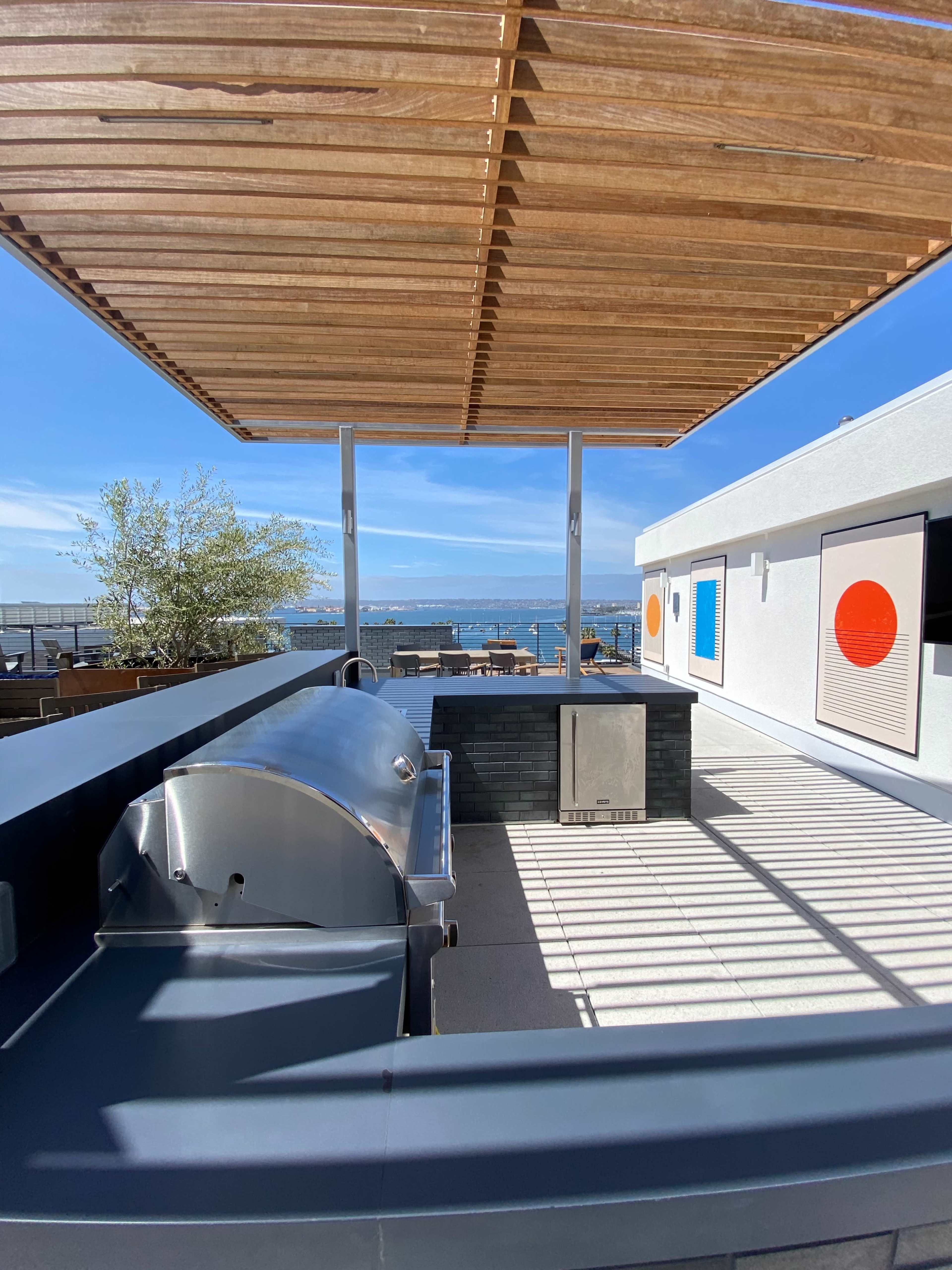The image shows a modern outdoor grilling area with a metal grill and a black stone counter, overlooking a body of water under a wooden pergola.