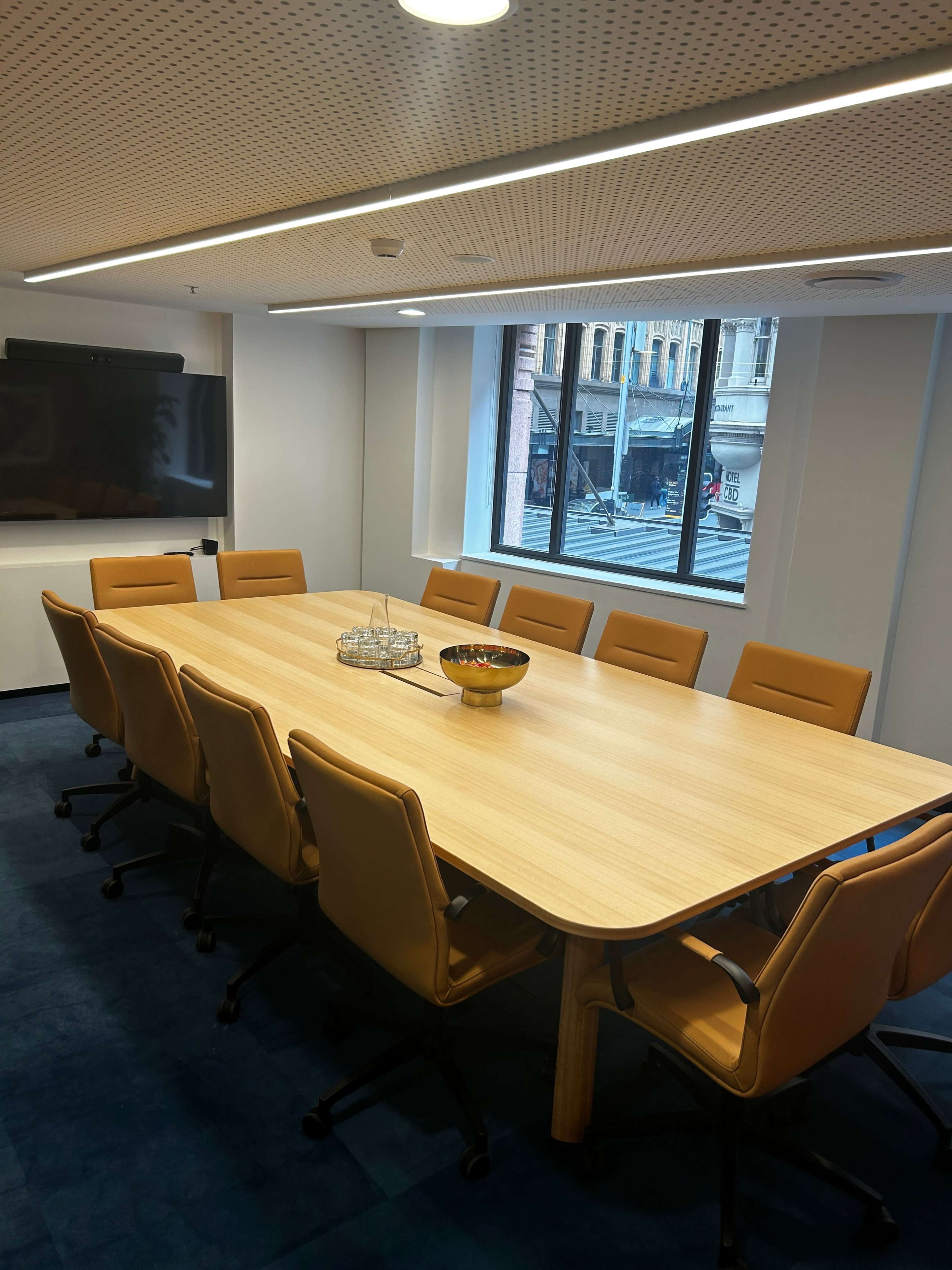A large conference room features a wooden table surrounded by ten orange chairs, with a bowl of fruit in the center and a view of the street through a window.
