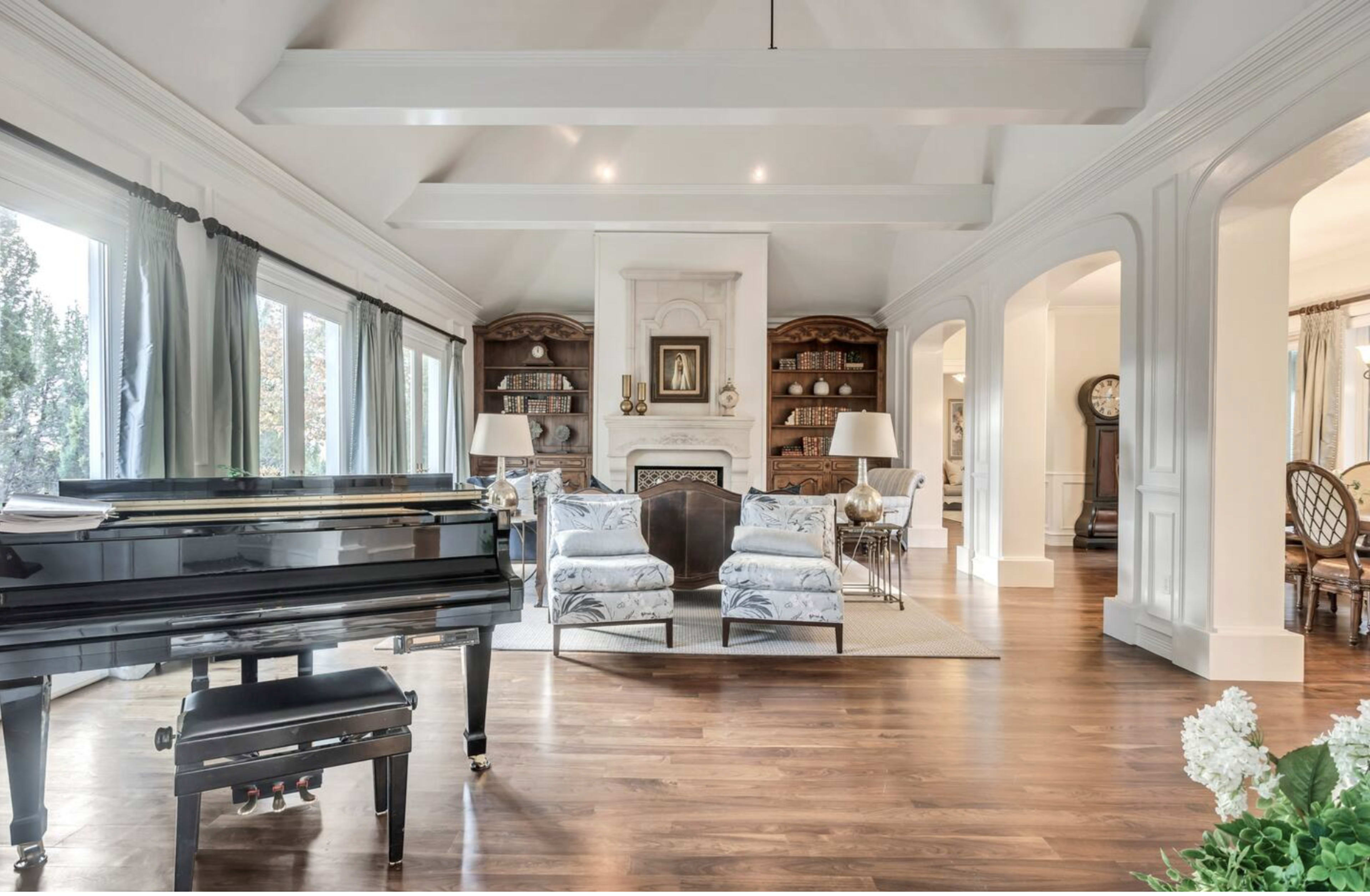 A spacious living area featuring a grand piano, two patterned armchairs, built-in bookshelves, and a dining area in the background.